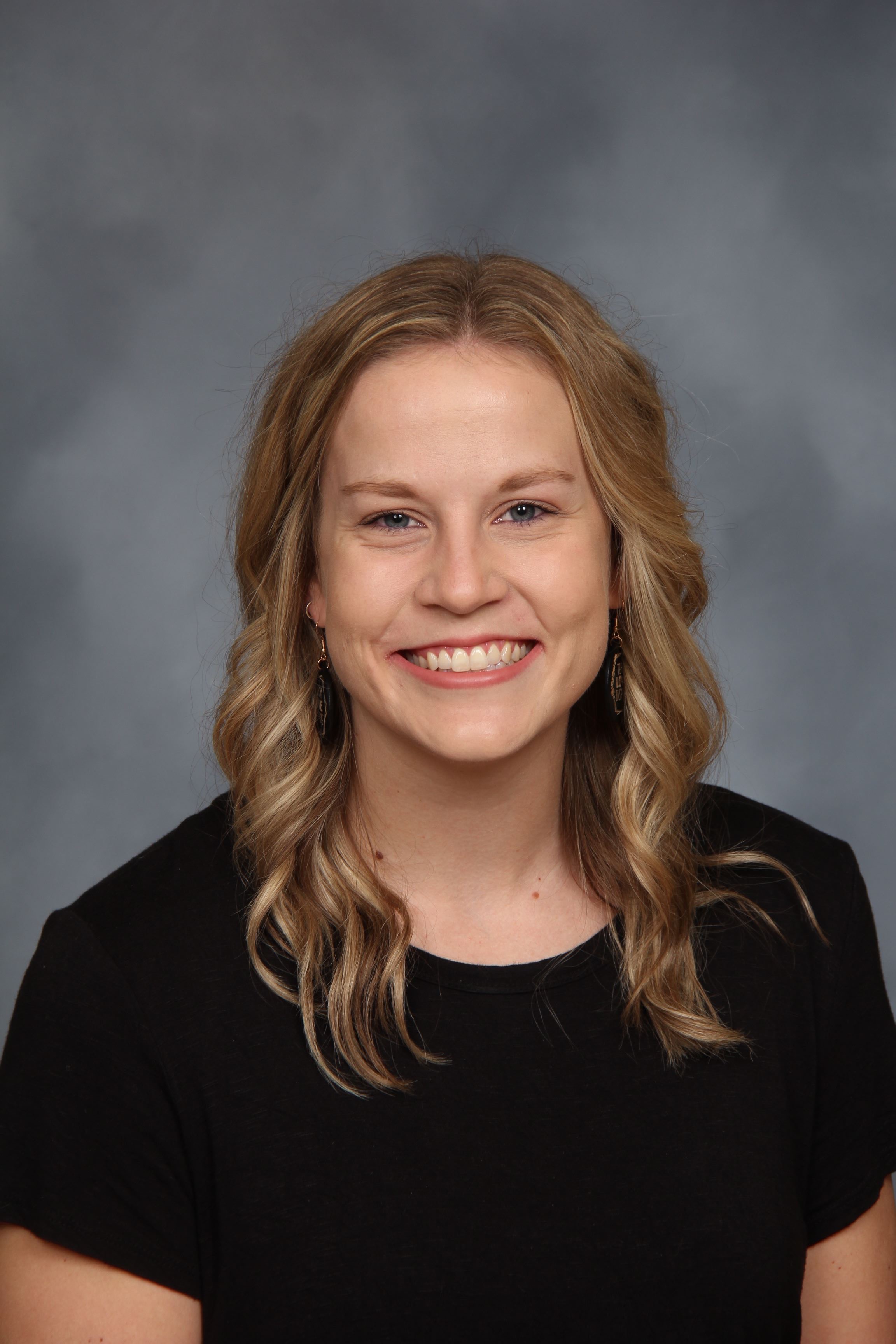 A woman with blonde hair, wearing a black shirt, smiles at the camera in front of a gray background.