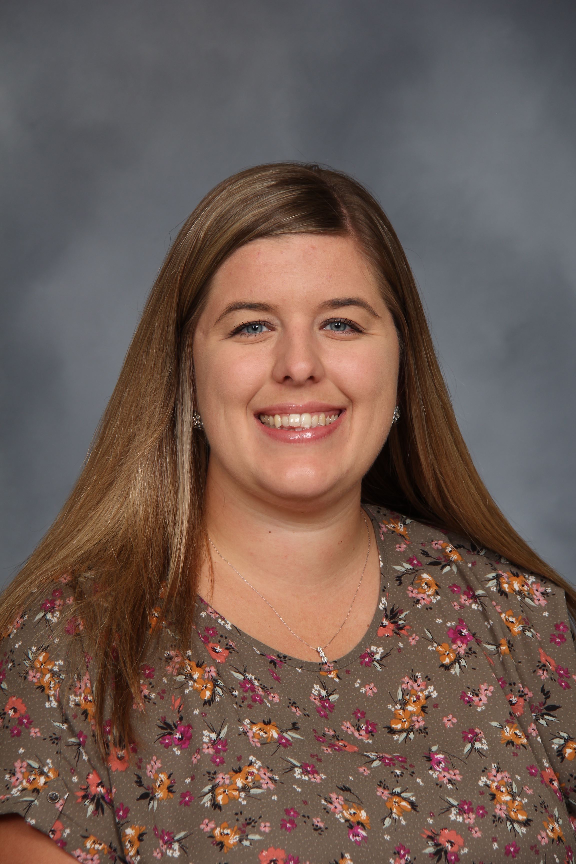 Woman with long hair smiles, wearing floral shirt, against a gray background.