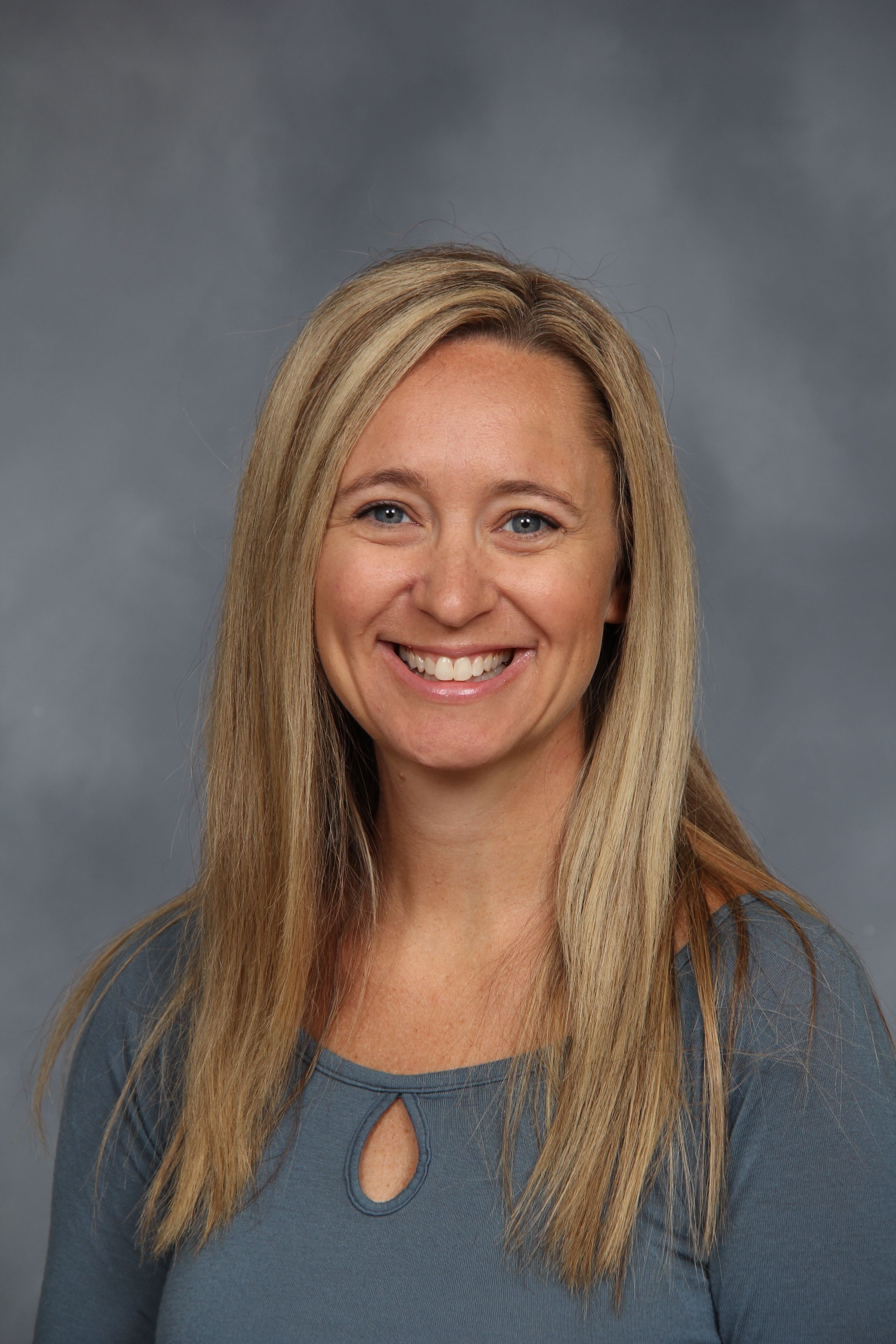 A woman with long blonde hair smiles at the camera in front of a gray background.