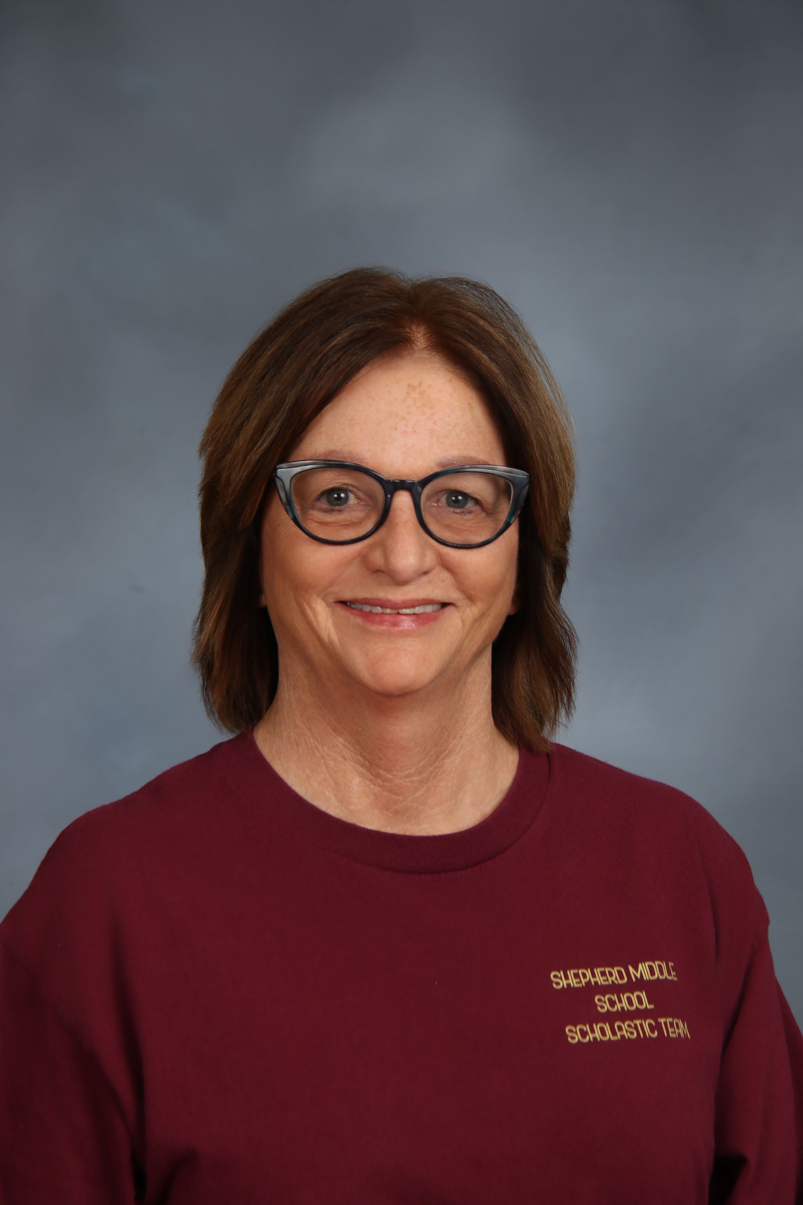Woman with short brown hair and glasses smiling, wearing a maroon shirt with white text.