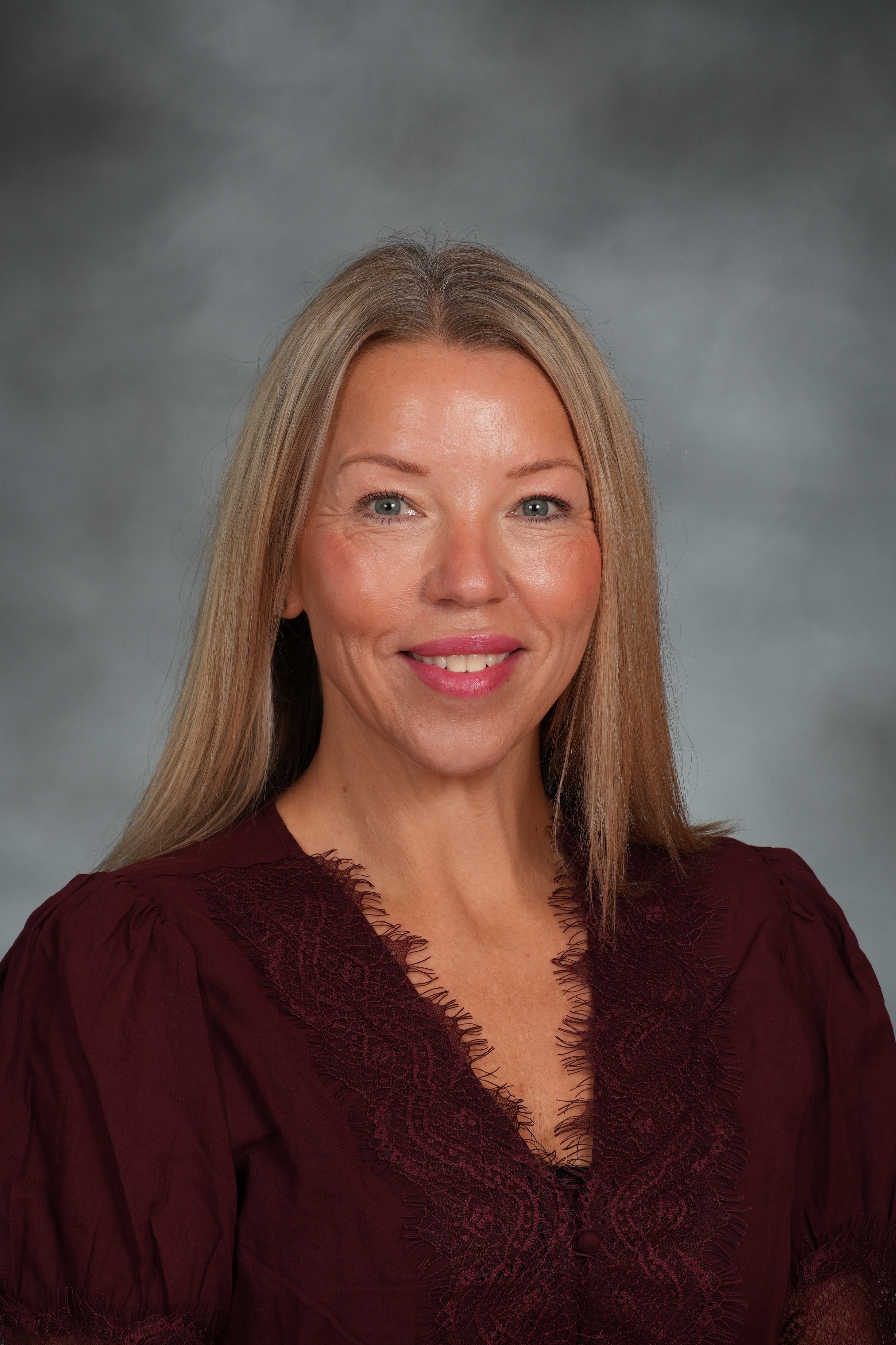 Woman with long, straight blonde hair, smiling, wearing a maroon blouse. Background is light gray.