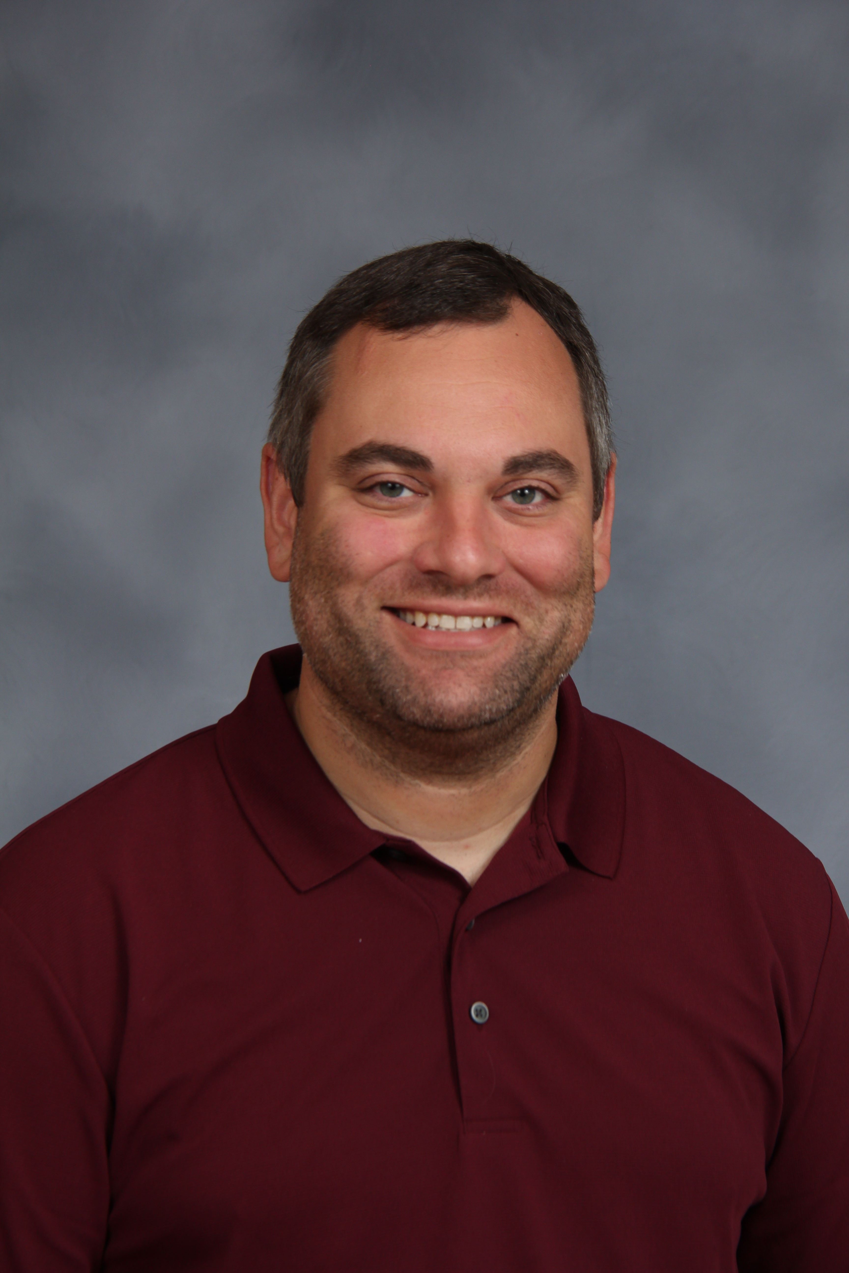 A smiling man with short hair in a maroon polo shirt. He is standing against a gray background.