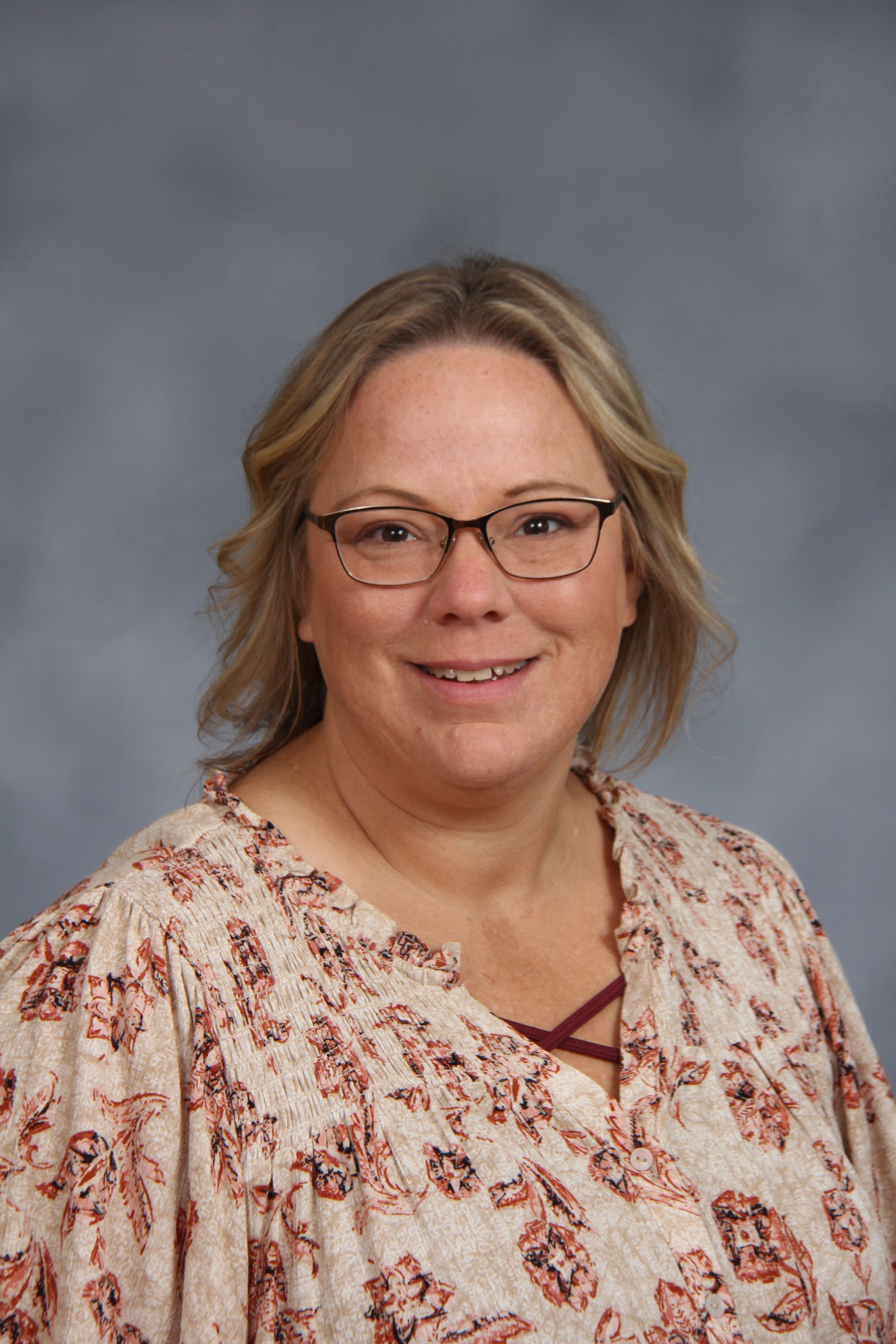 A woman with short blonde hair and glasses smiles at the camera. She wears a floral blouse.