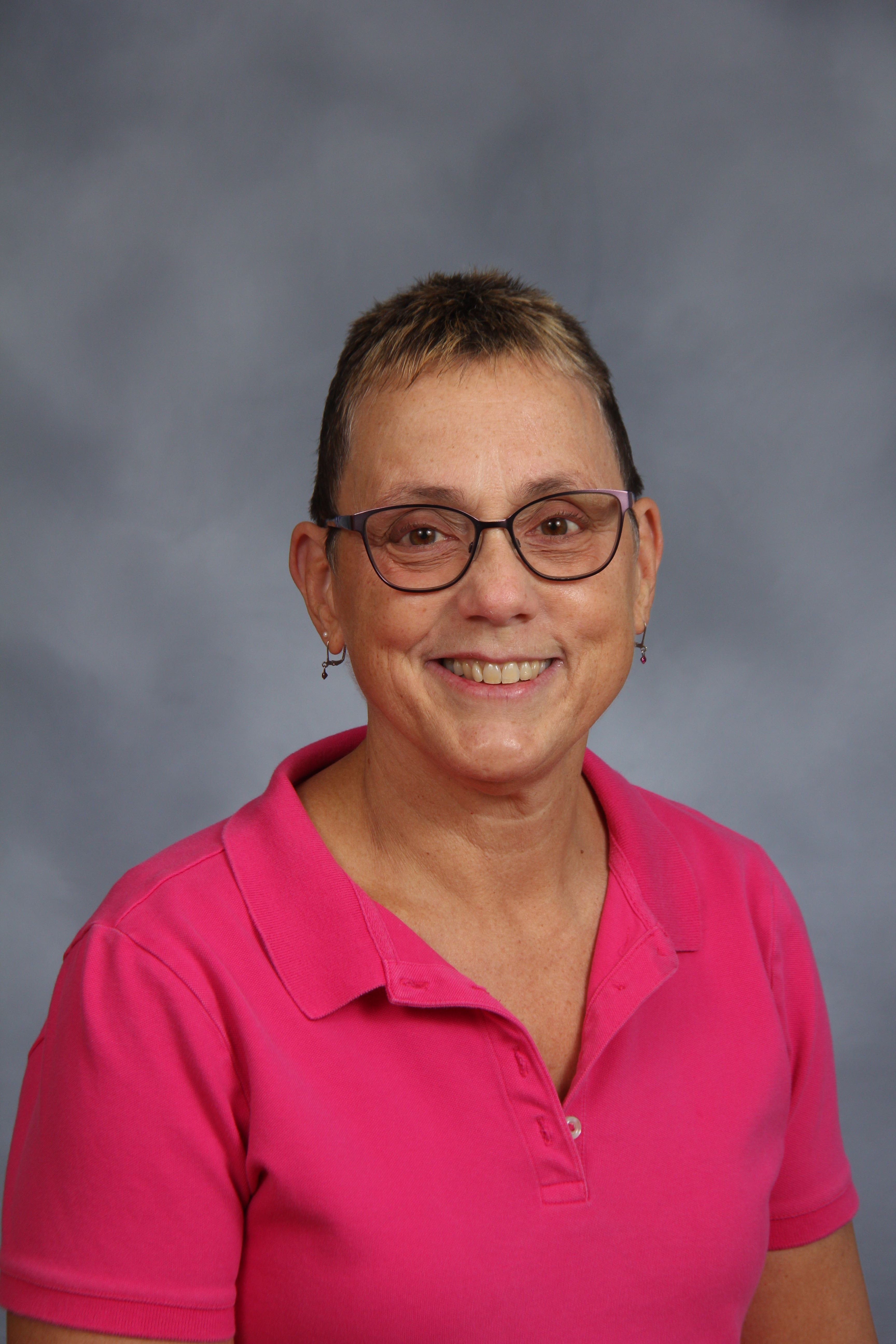 A woman wearing glasses and a pink polo, smiling at the camera, stands against a gray background.