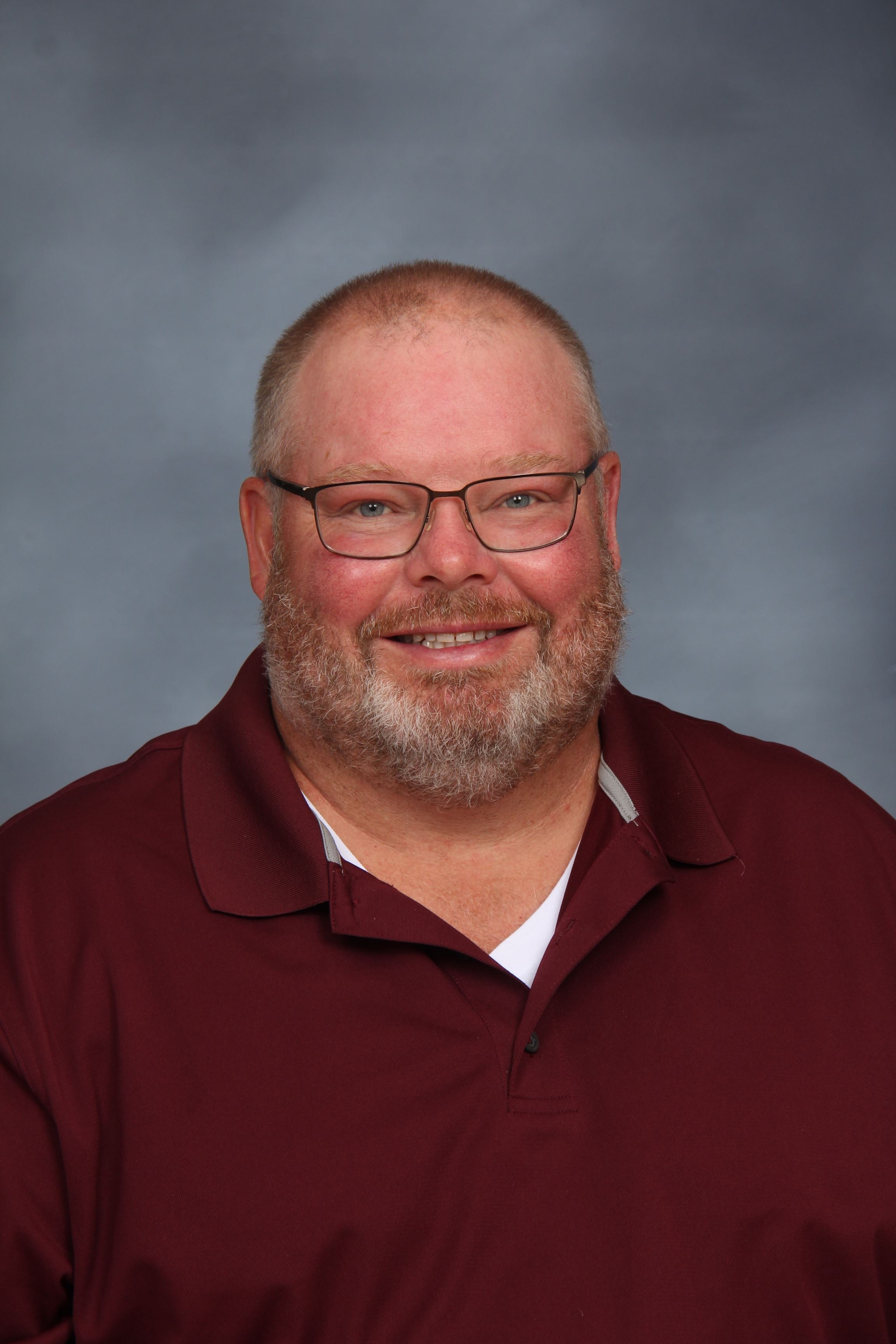 A man with a beard and glasses smiles at the camera. He wears a maroon polo shirt. The background is gray.