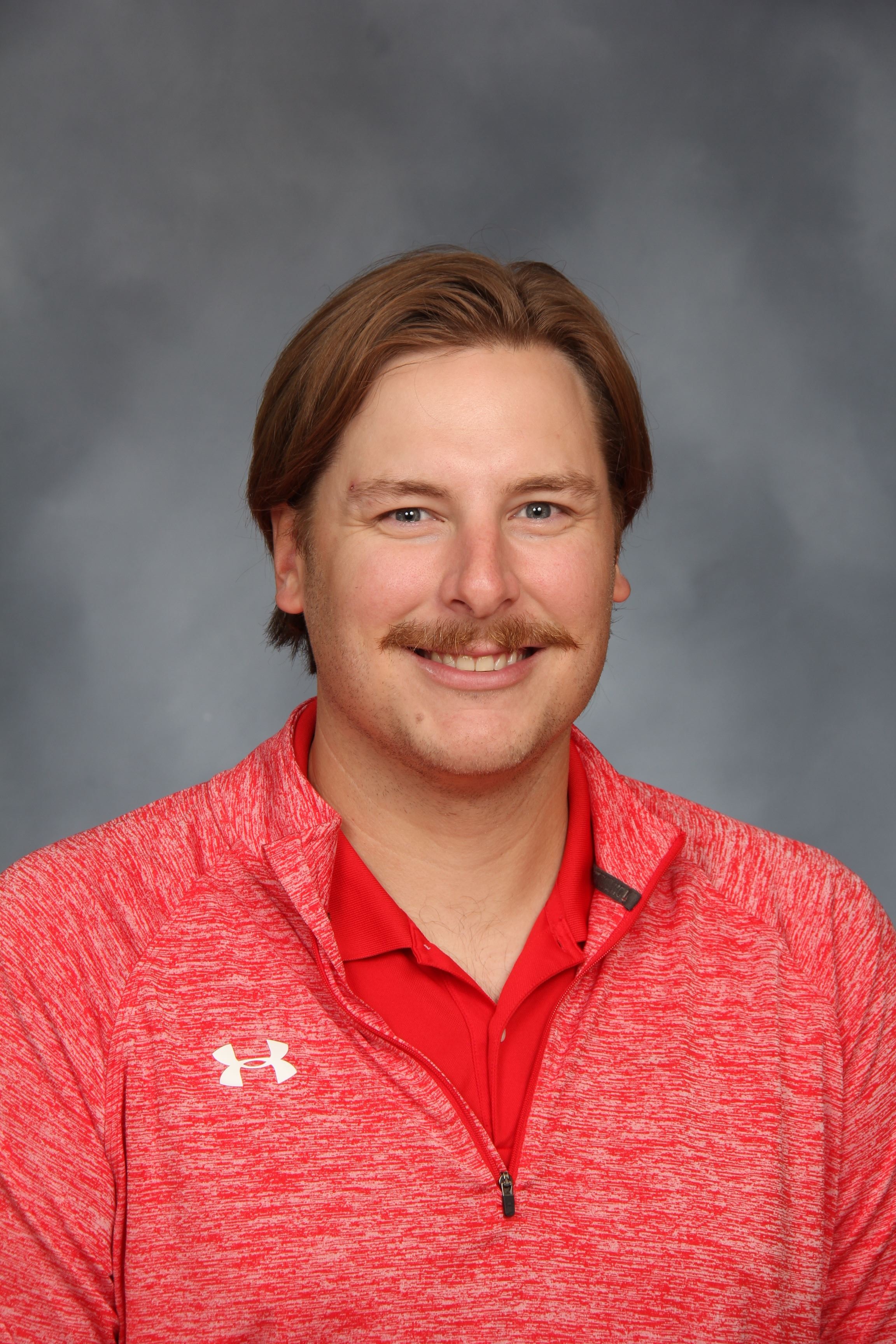 A man with short hair and a mustache in a red shirt with a small logo. He is smiling.