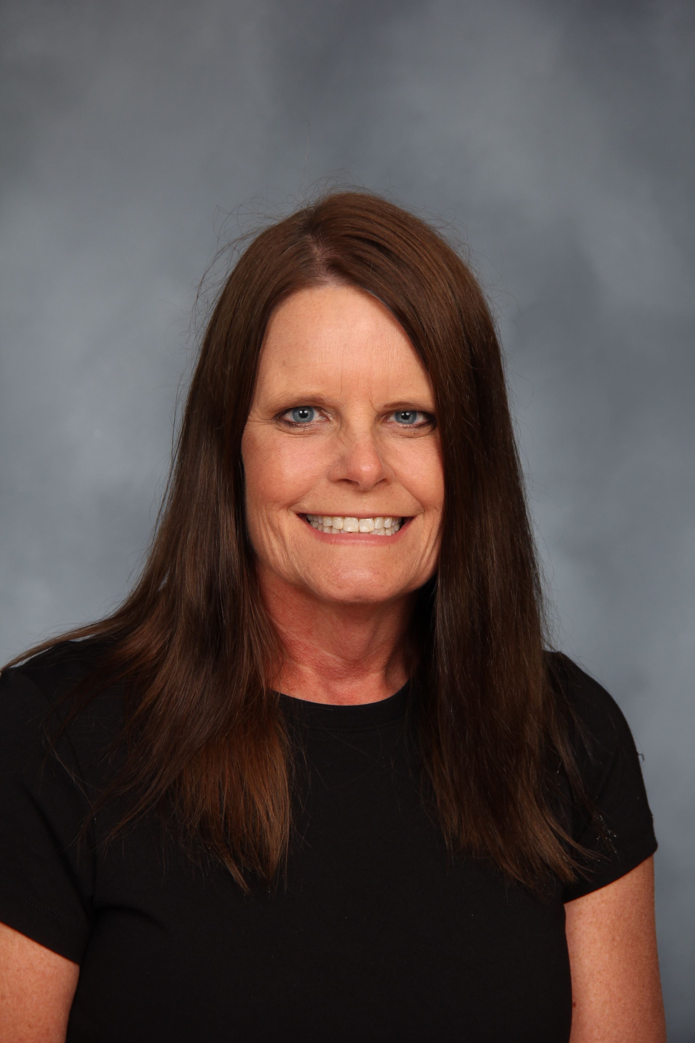 A woman with long brown hair smiles, wearing a black shirt. Background is gray.