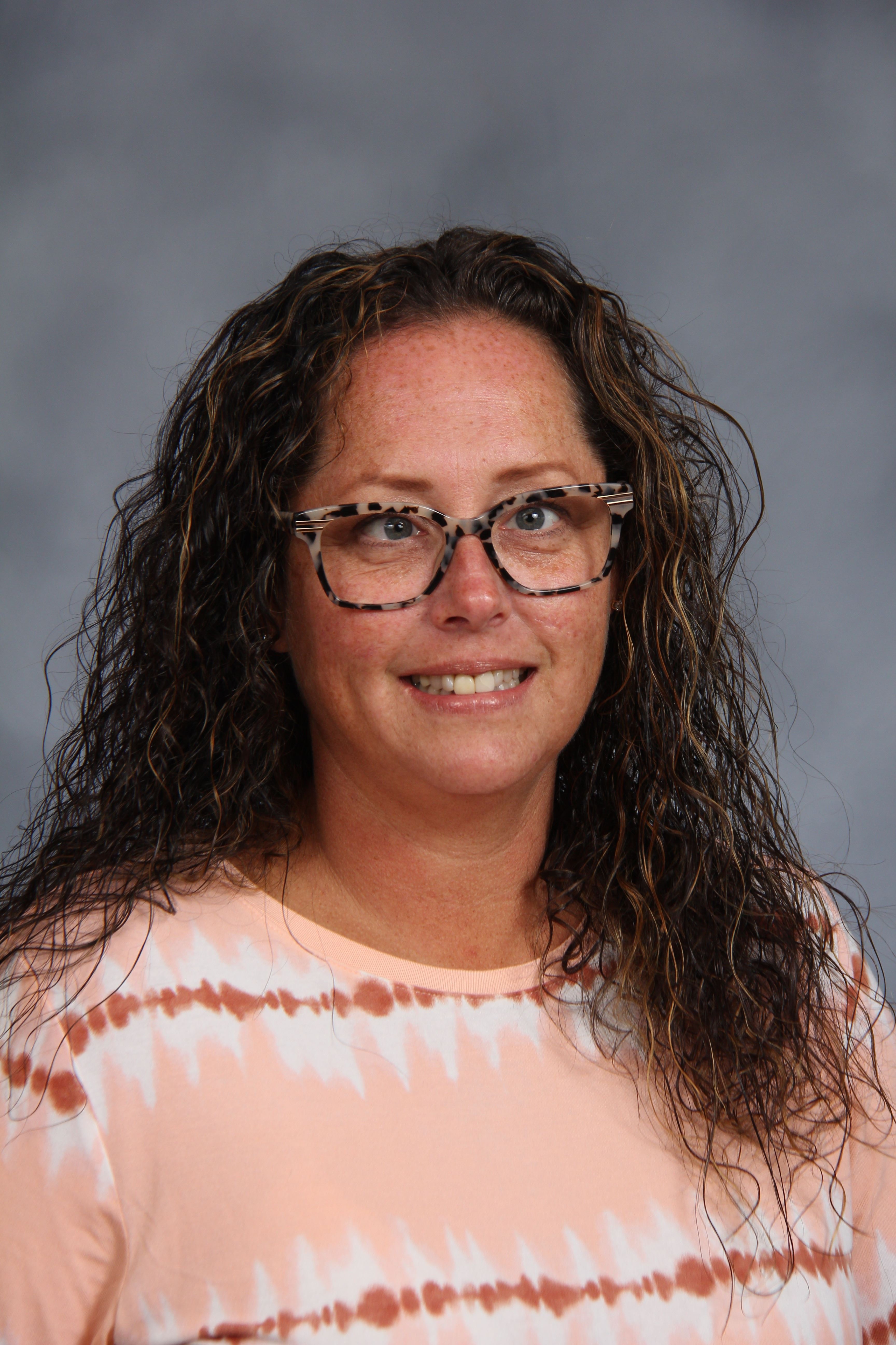 Woman with long curly hair and glasses. Wearing a pink tie-dye shirt. Smiling. Background is gray.