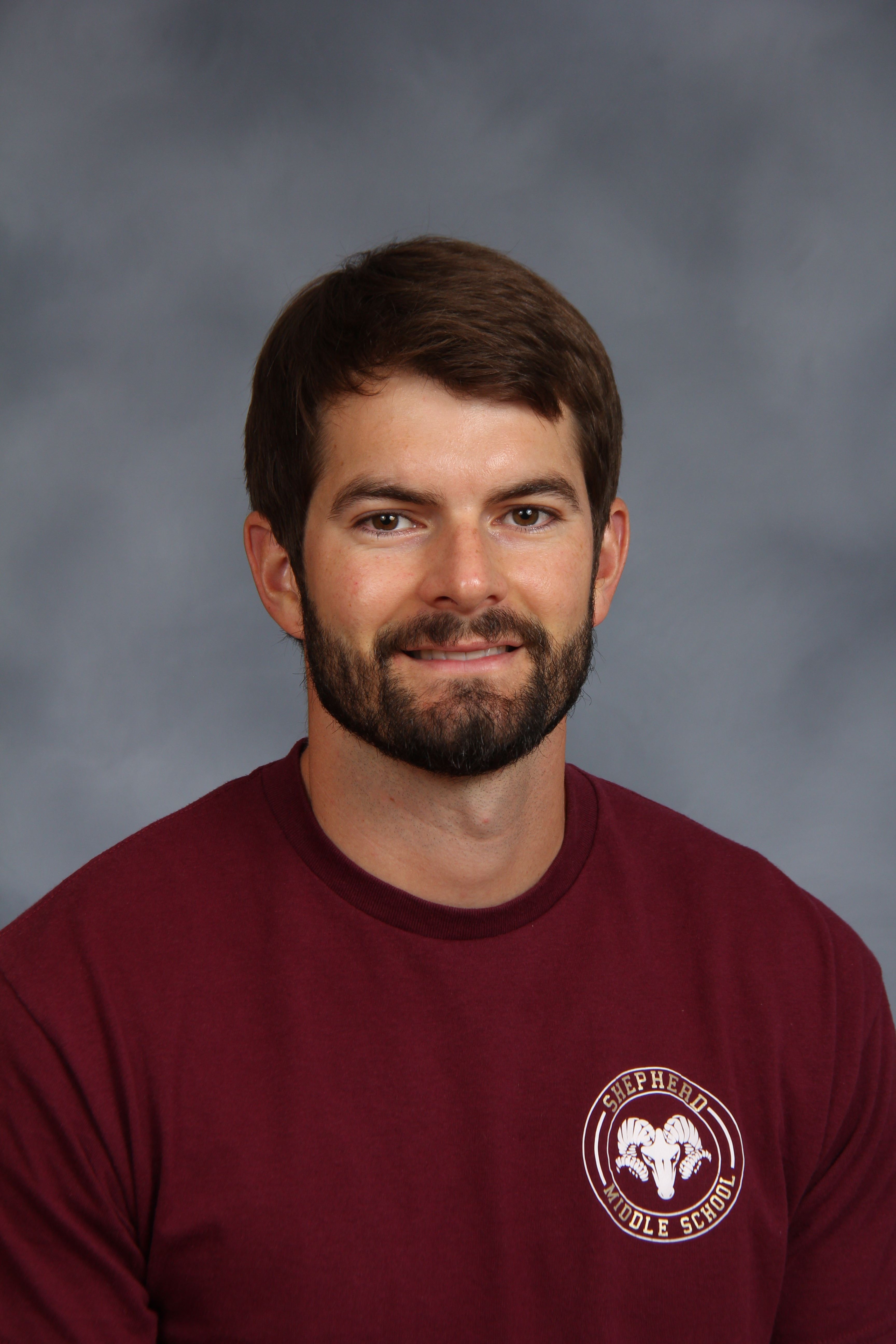 Man in maroon shirt with a logo on the left chest, standing in front of a gray background.