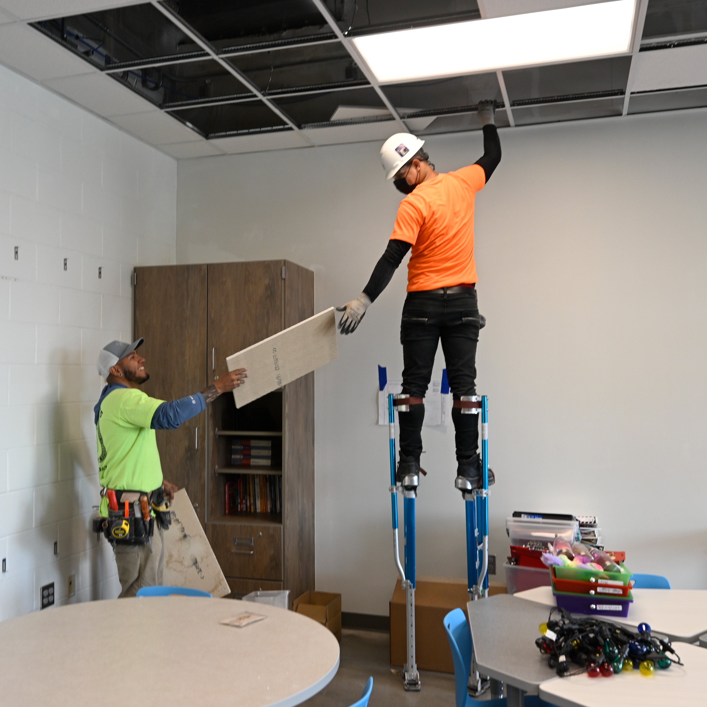Man handing another man on stilts ceiling tile to install