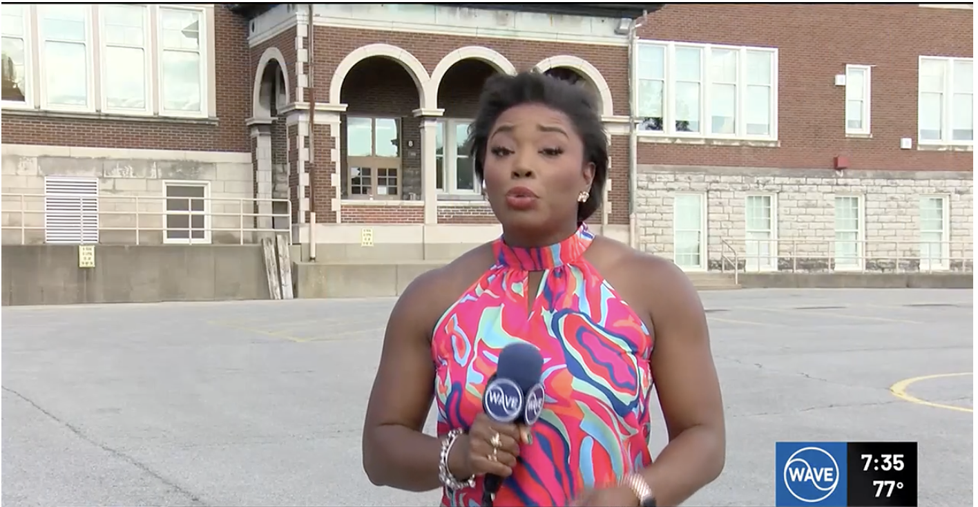 A journalist holds a microphone in front of Breck Metro High School. 