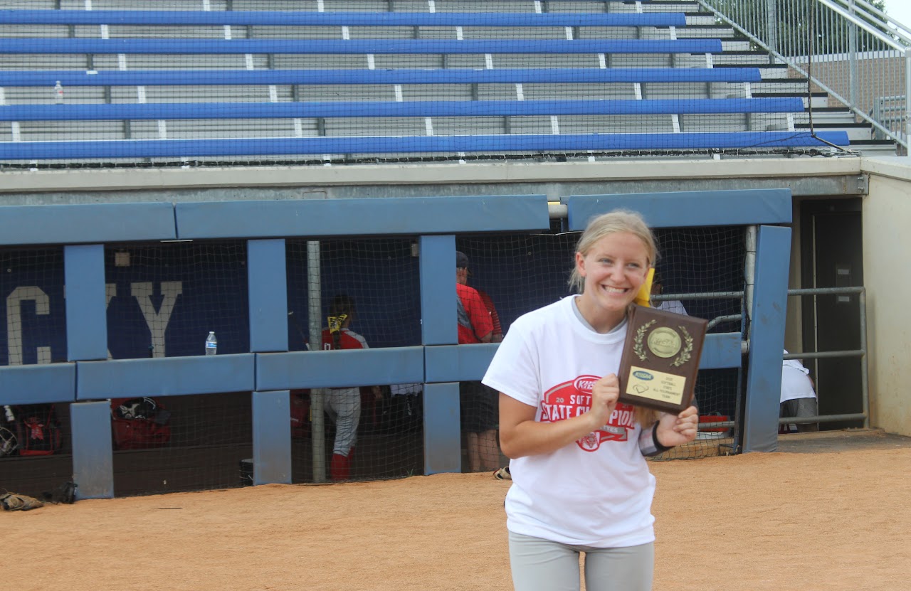 Young girl posing with trophy in front of baseball dugout.