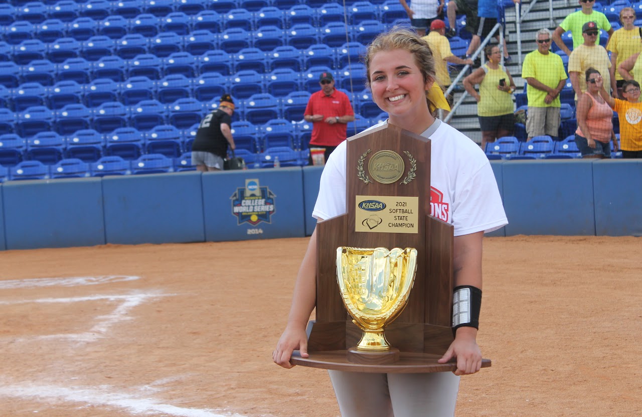 Young girl posing with trophy in front of baseball dugout.