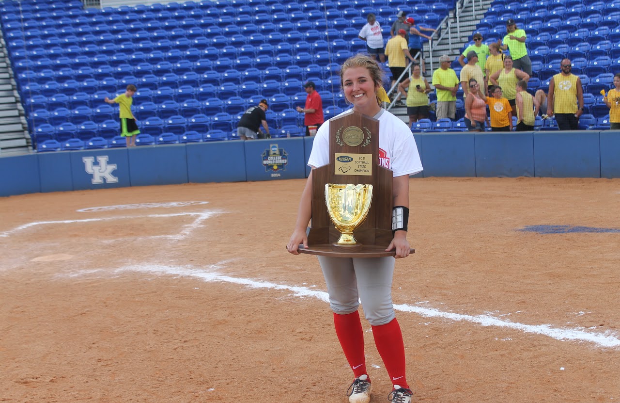 Young girl posing with trophy in front of baseball dugout.