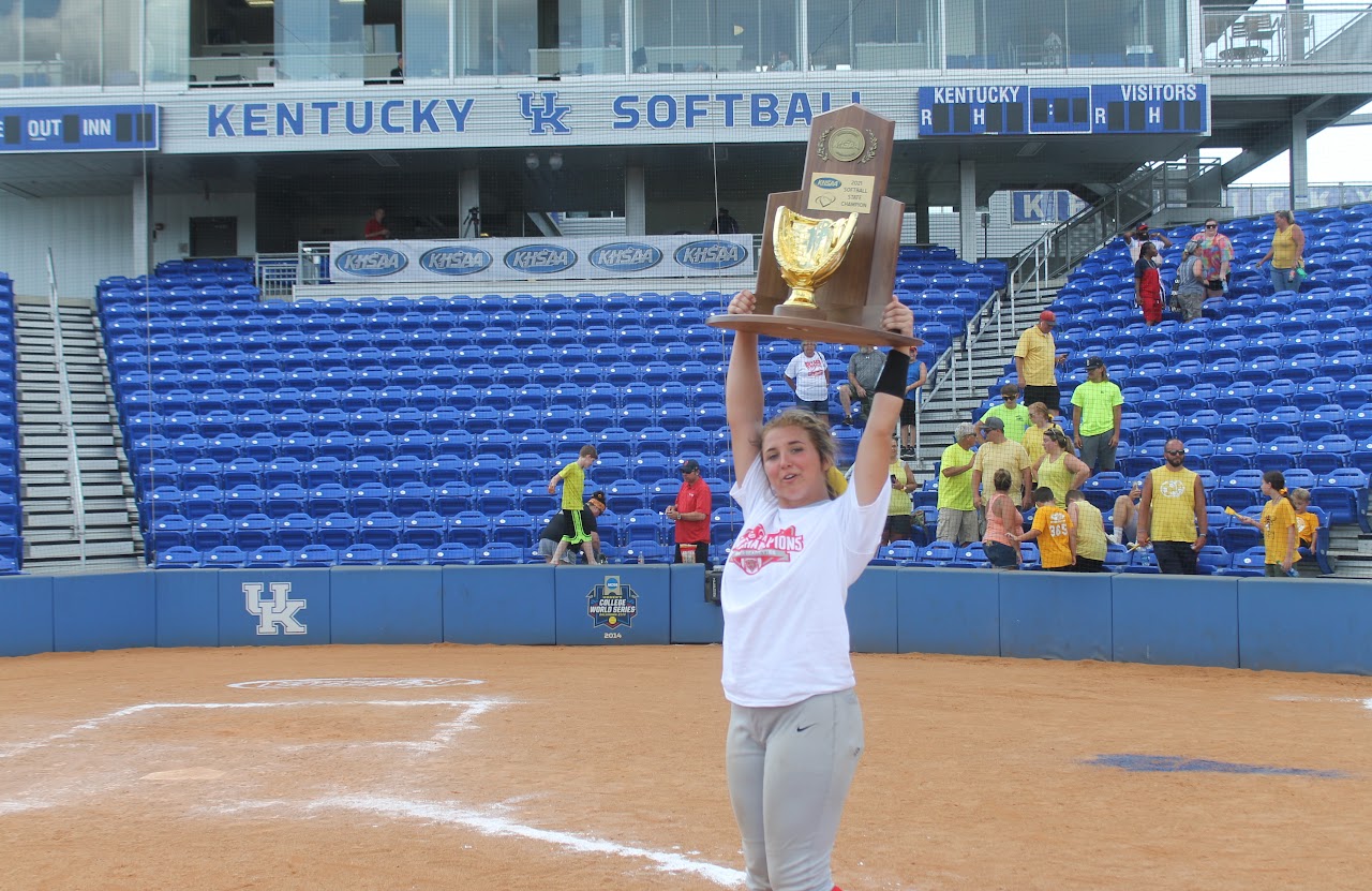 Young girl posing with trophy in front of baseball dugout.