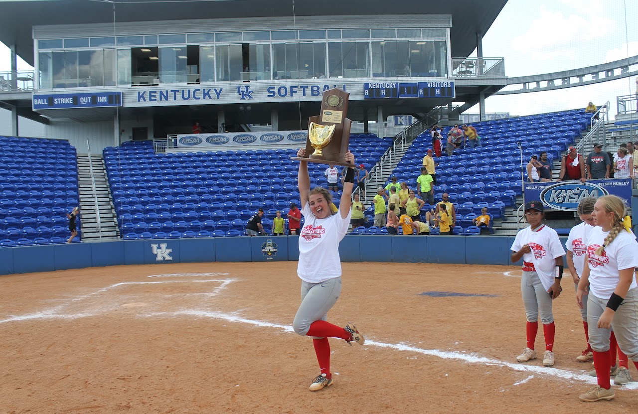 Young girl posing with trophy in front of baseball dugout.