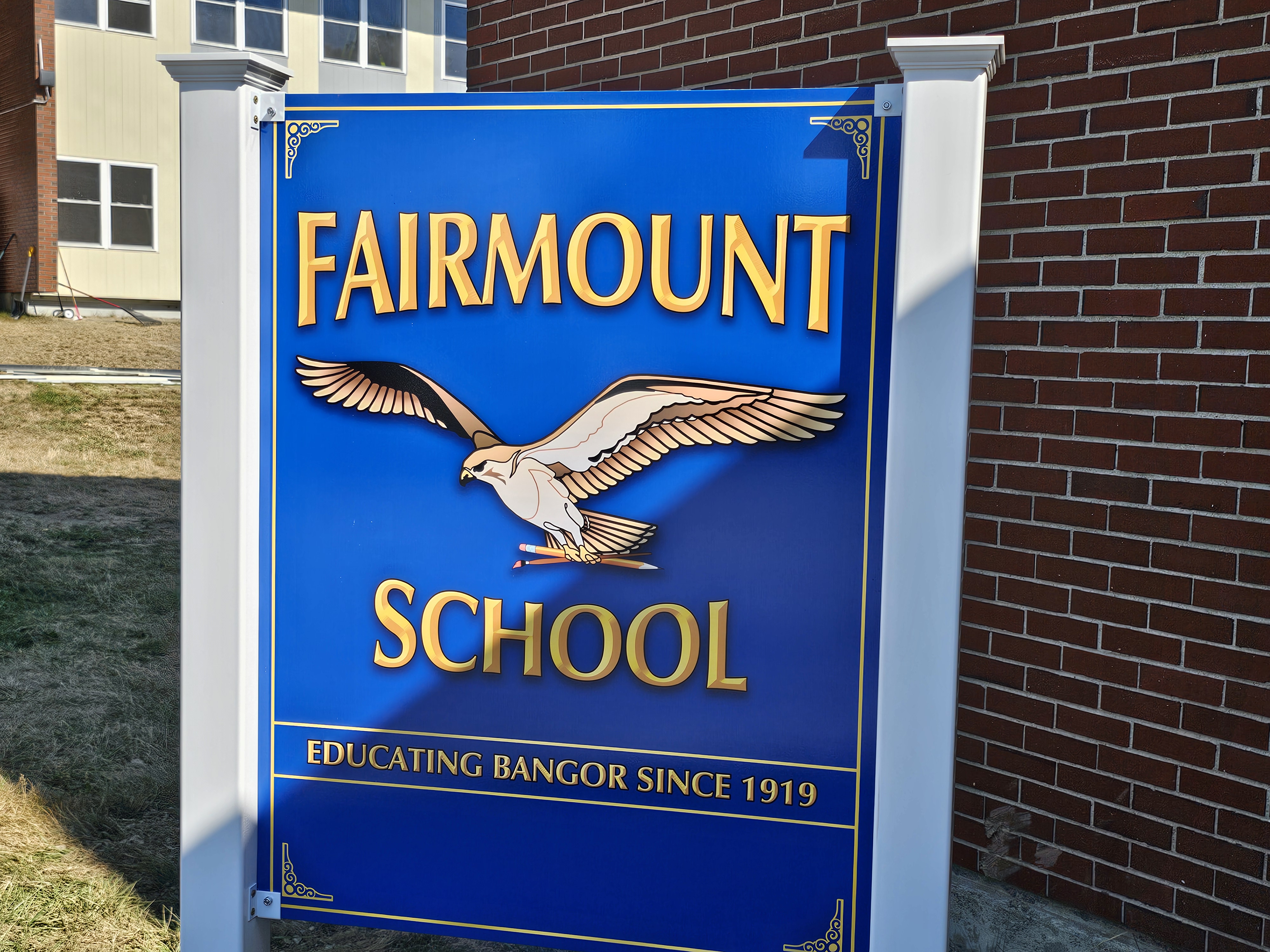 Blue sign with a bird on it. The text reads "Fairmount School" and "Educating Bangor Since 1919". Brick building in background.