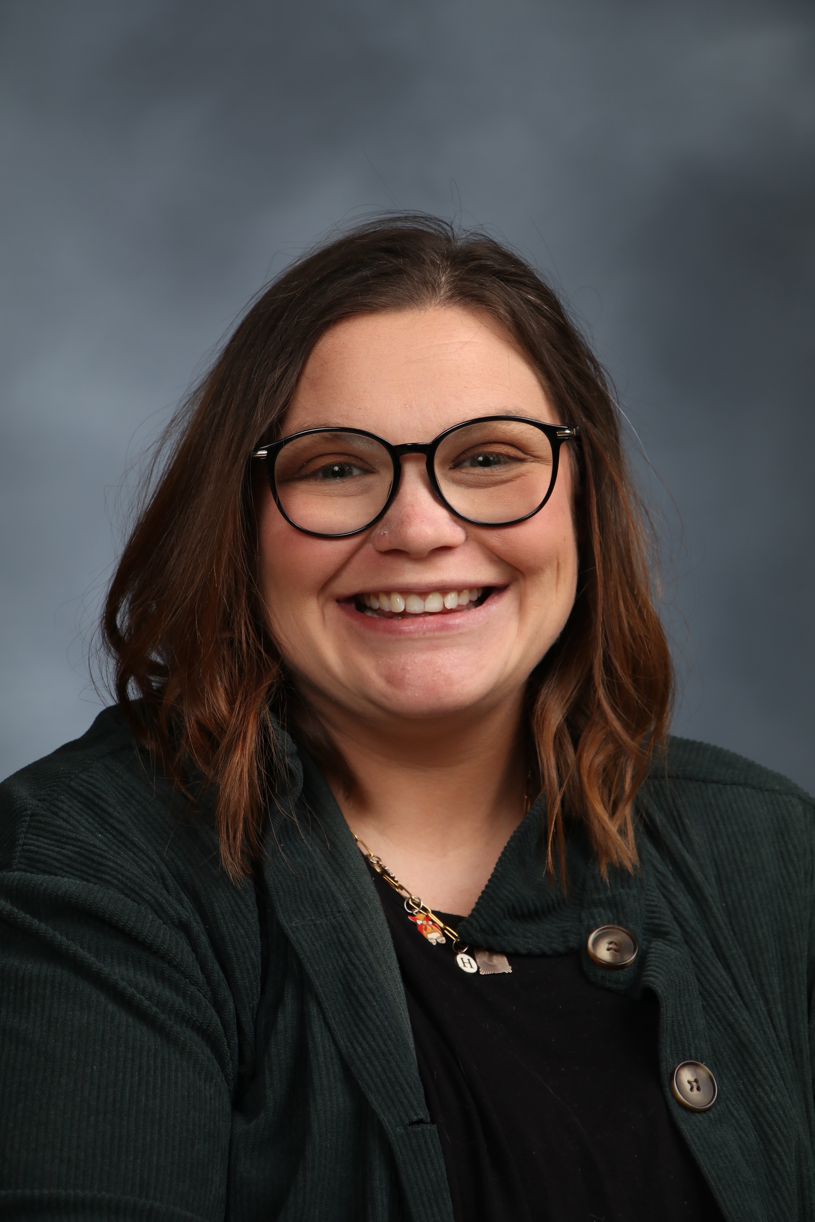 A woman wearing glasses and a green cardigan over a black top, smiling against a solid gray background.