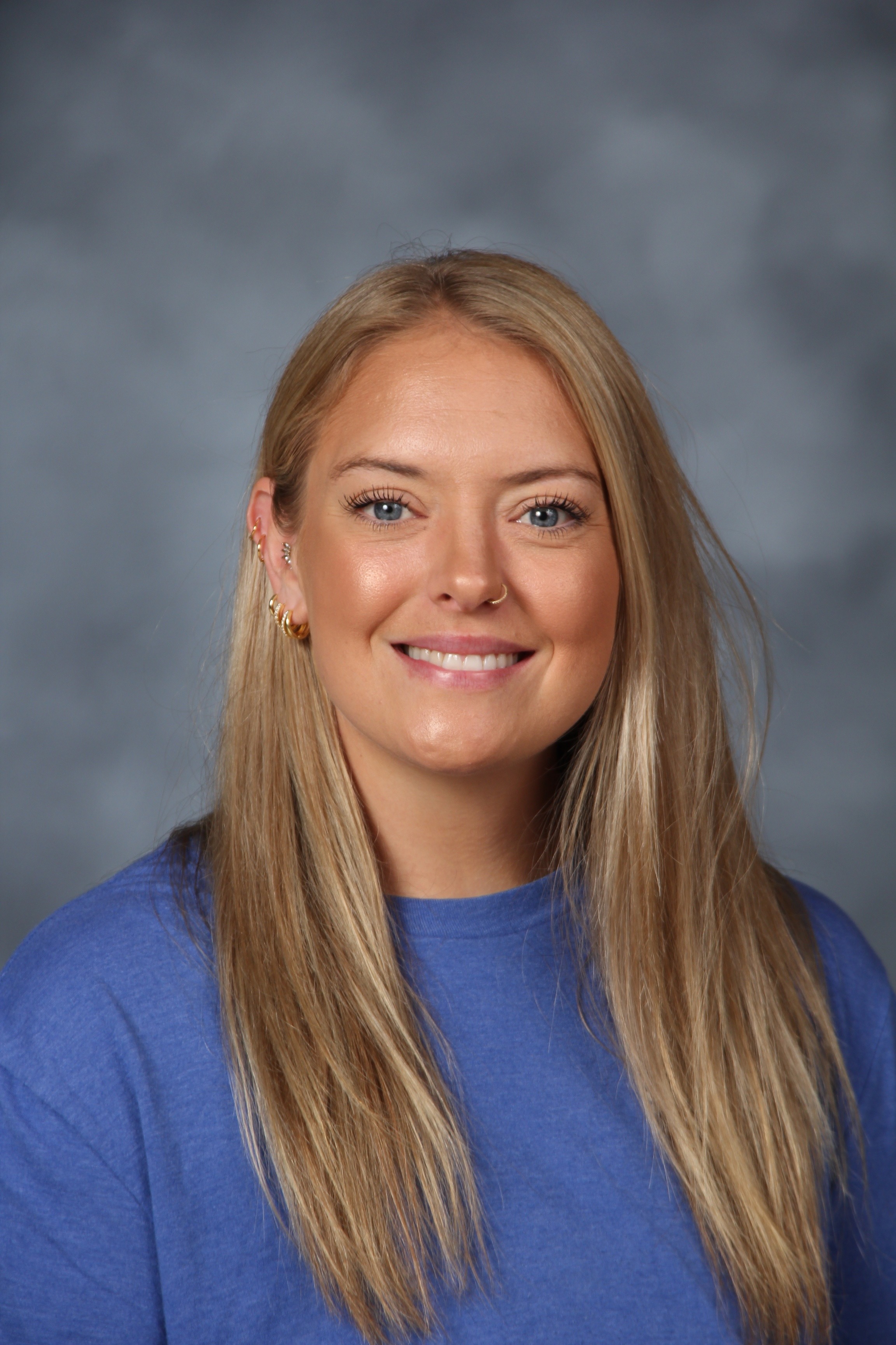 Woman with blonde hair in a blue shirt, smiling, standing in front of a gray background.
