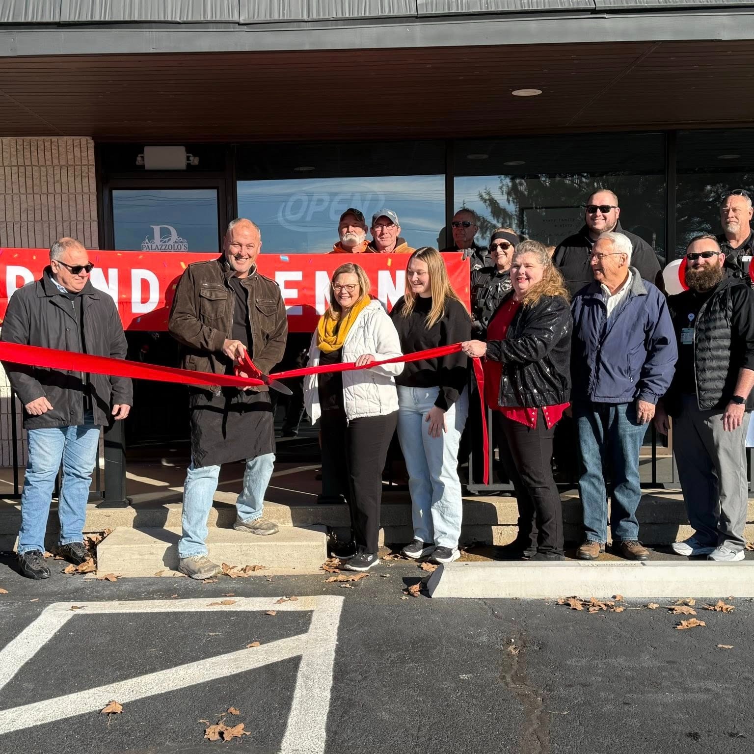 The grand opening day at Palazzolo's Italian Restaurant and Pizzeria on HWY FF. A man in a brown jacket cuts a red ribbon.