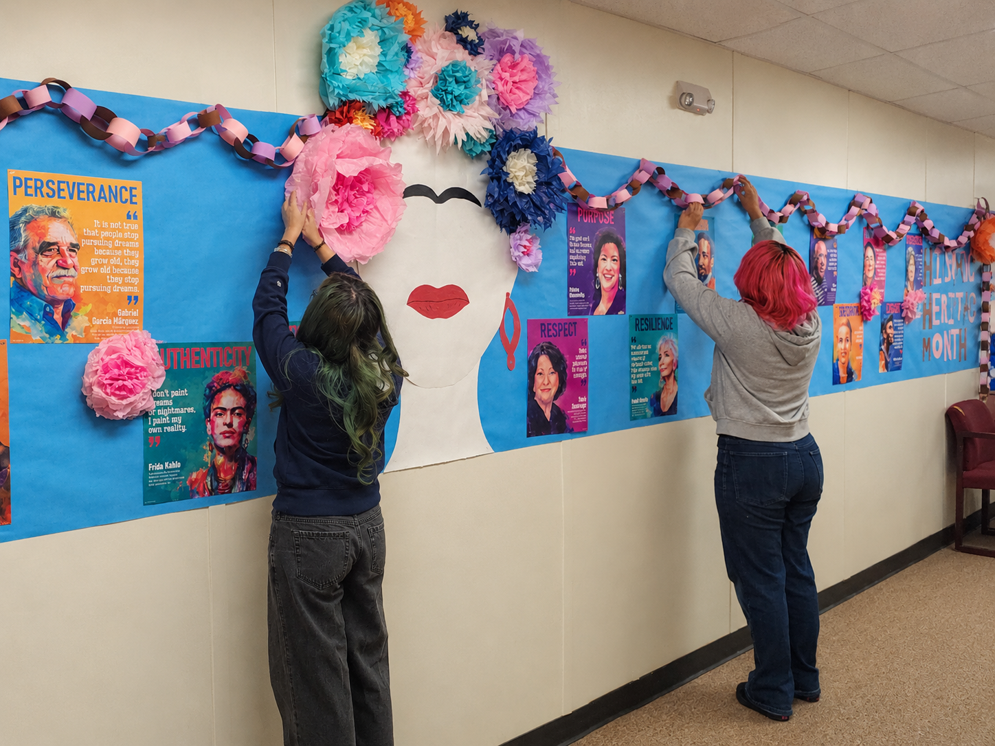 Two people decorating a wall with a Frida Kahlo portrait, flowers, and text.