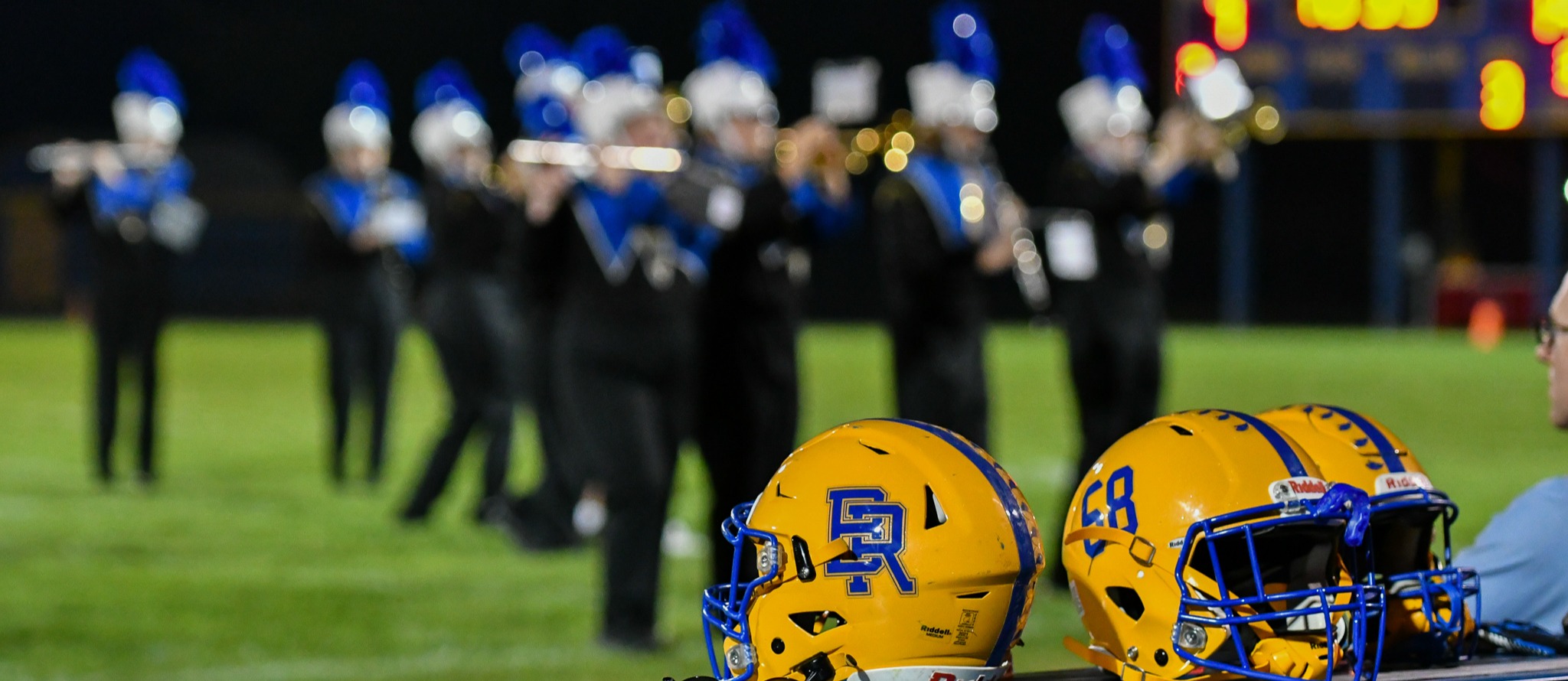 A Birch Run football helmet sits on a bench in front of the band playing on the field.