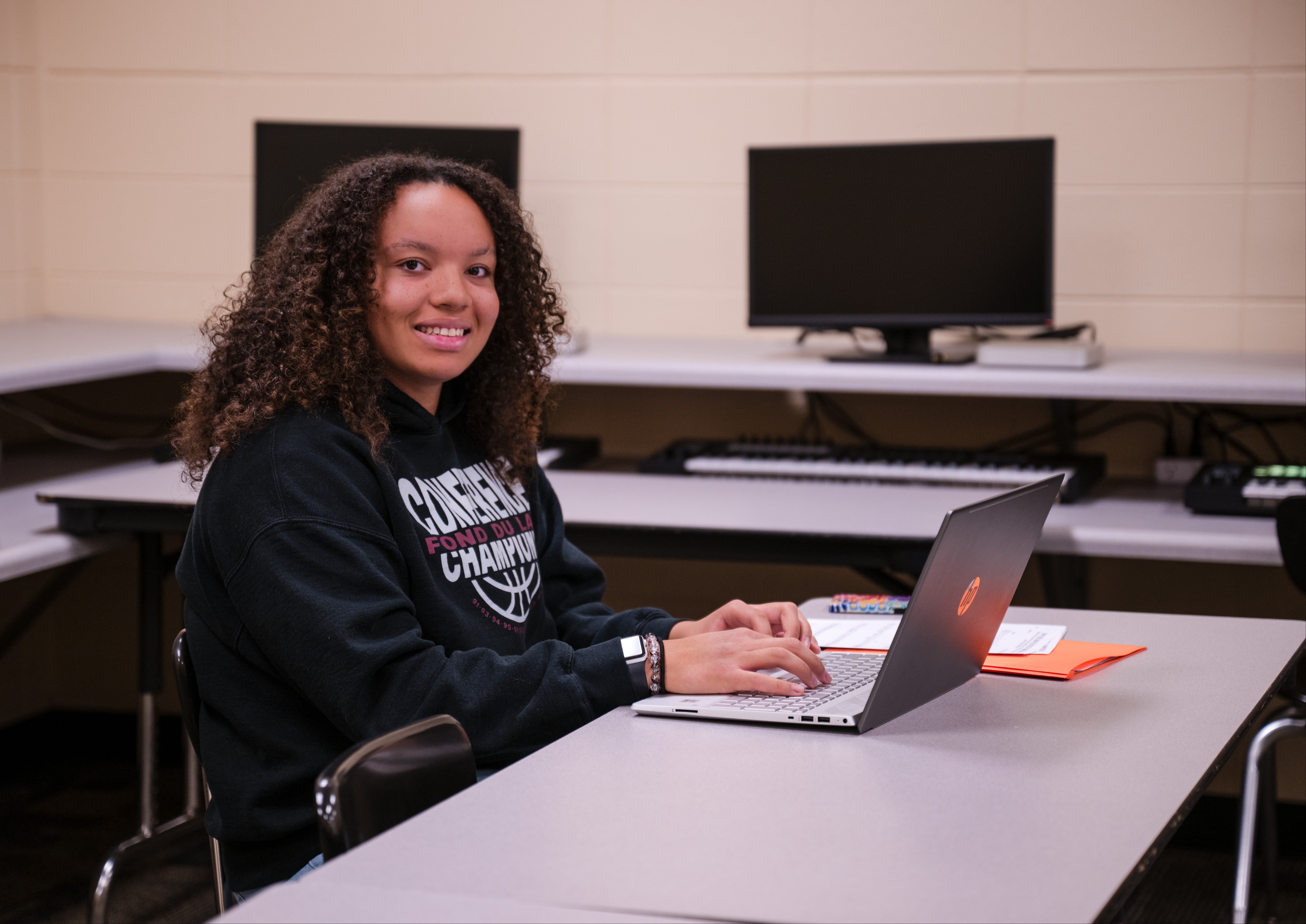 Senior student Amaria wearing a Fond du Lac Conference Champions basketball sweatshirt smiling at the camera while working on a laptop