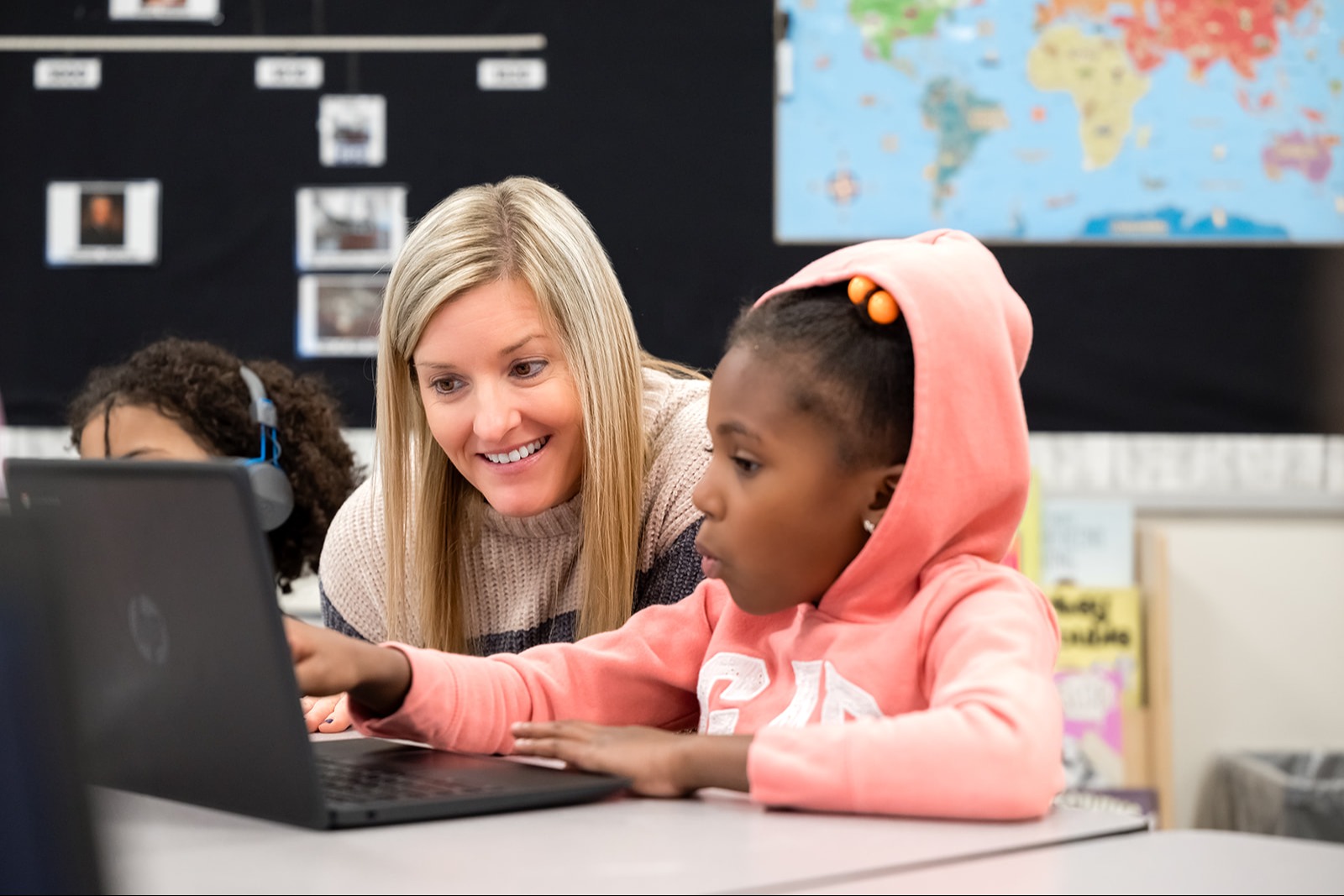  Principal Hughes smiles while assisting a student in a pink hoodie with a laptop in a classroom.