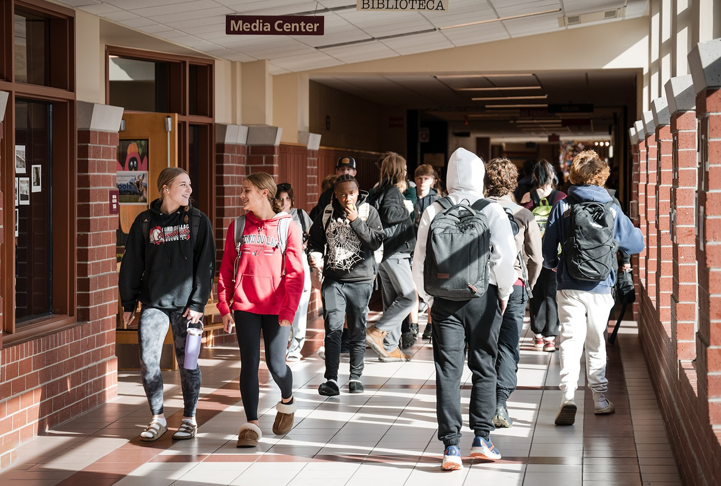 A brightly lit, interior hallway of the hight school with large windows. There at students walking to and from class.