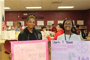 Two young students in a library, each holding up a poster board displaying research findings.