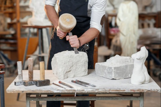 A person in a studio holds a mallet, carving a stone sculpture. Tools and partial sculptures are on the table.