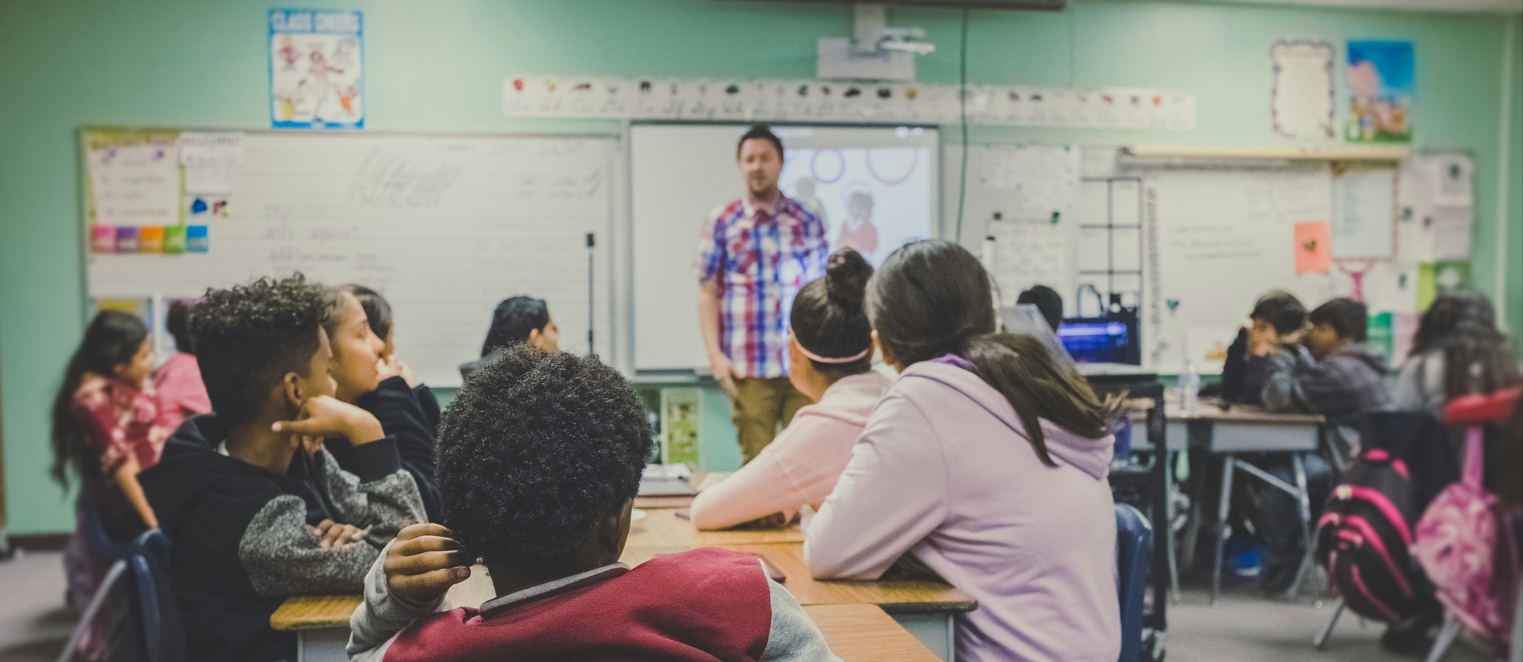 A classroom with a teacher at a podium in front of students seated at desks, listening.
