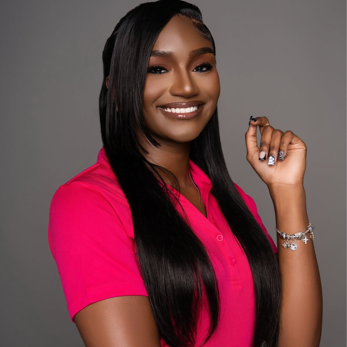 A smiling woman with long black hair wearing a pink shirt poses against a gray background. She has manicured nails.