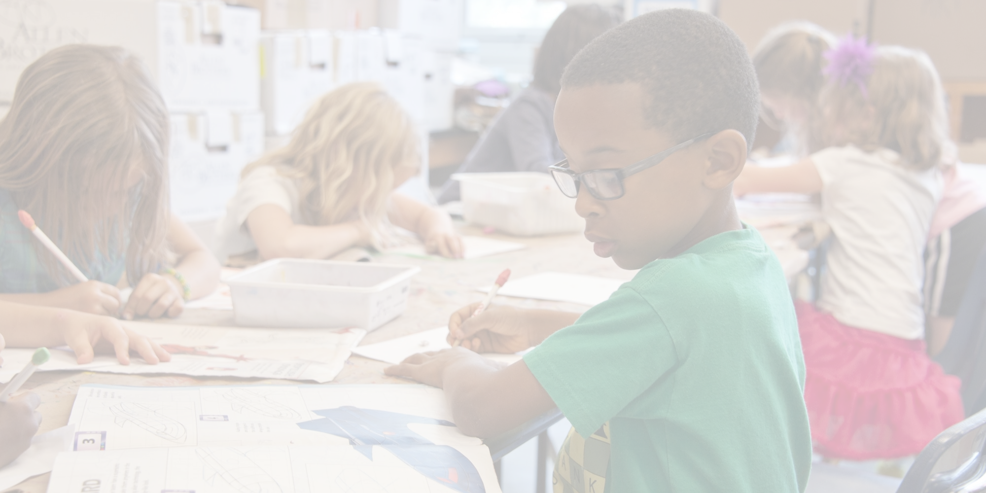 Several young children in a classroom sit at desks. One child wears glasses and writes with a pencil.