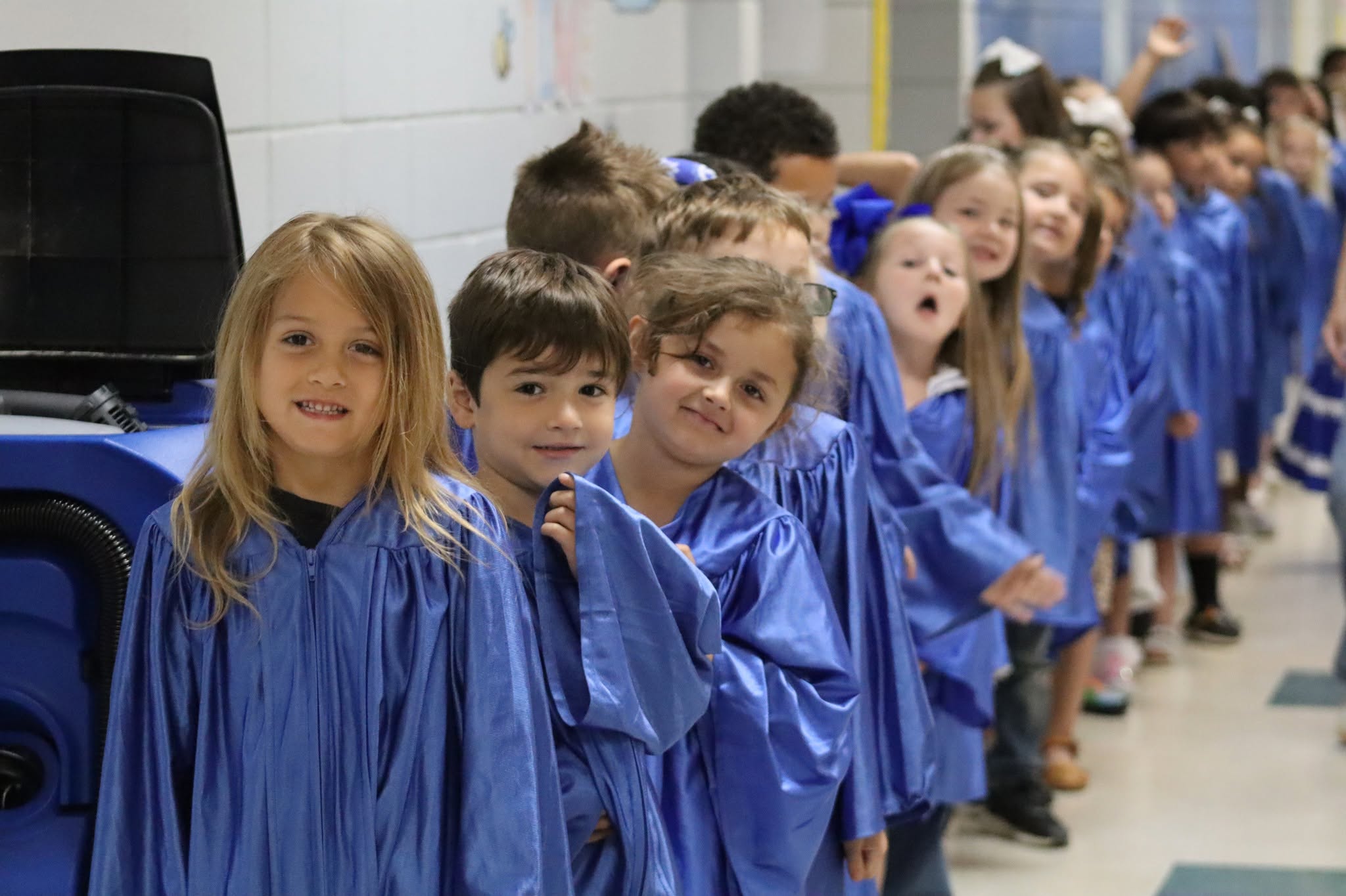 A row of children in blue gowns, smiling and standing in a school hallway.