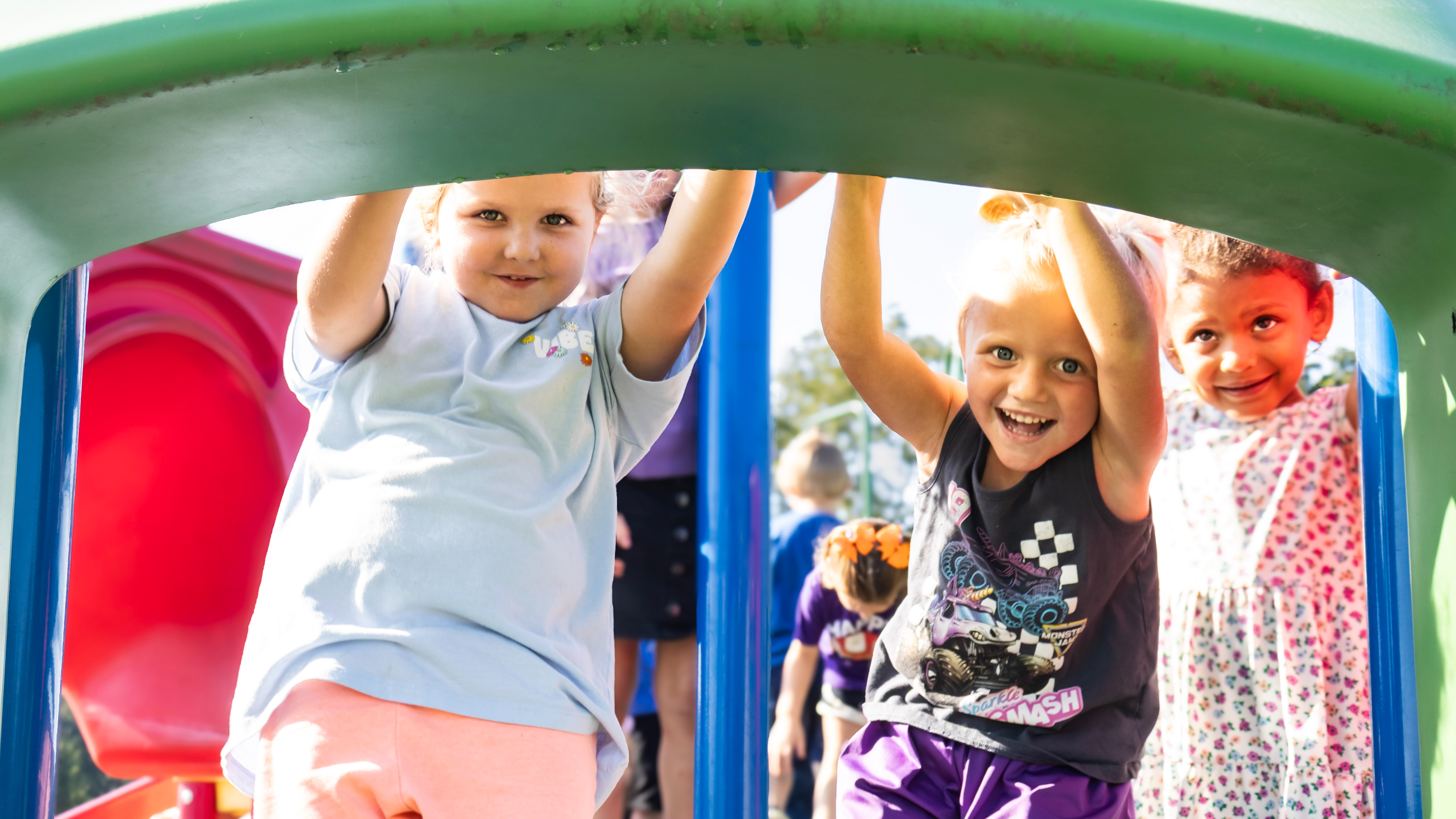 Three young children play in a playground. They are holding onto a green arch, with others in the background.