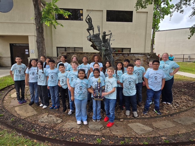 A group of children in blue shirts pose with a trophy in front of a building.
