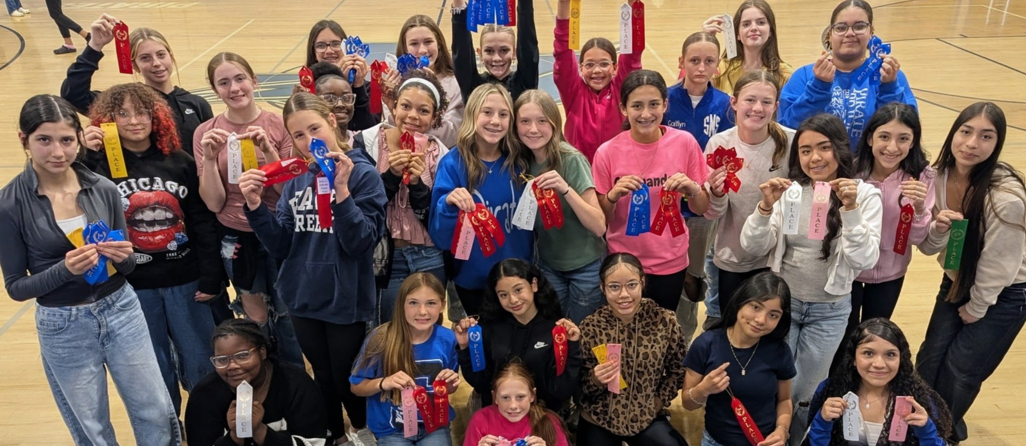 A group of students posing for a photo holding red ribbons. Some wear sneakers, and others wear jeans. Background includes a basketball court.