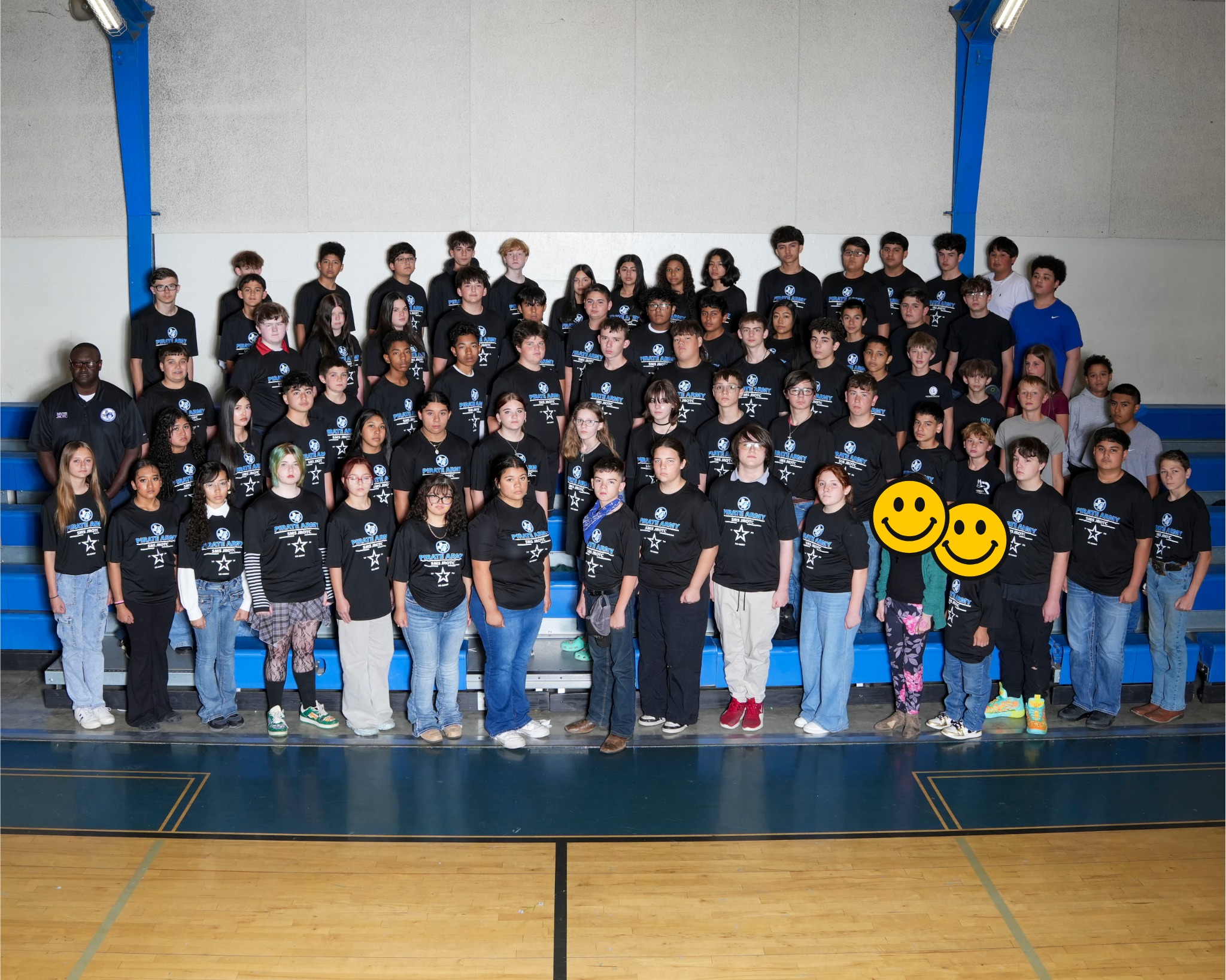 Large group in black t-shirts, smiling, posing for a photo in a gymnasium with blue bleachers.