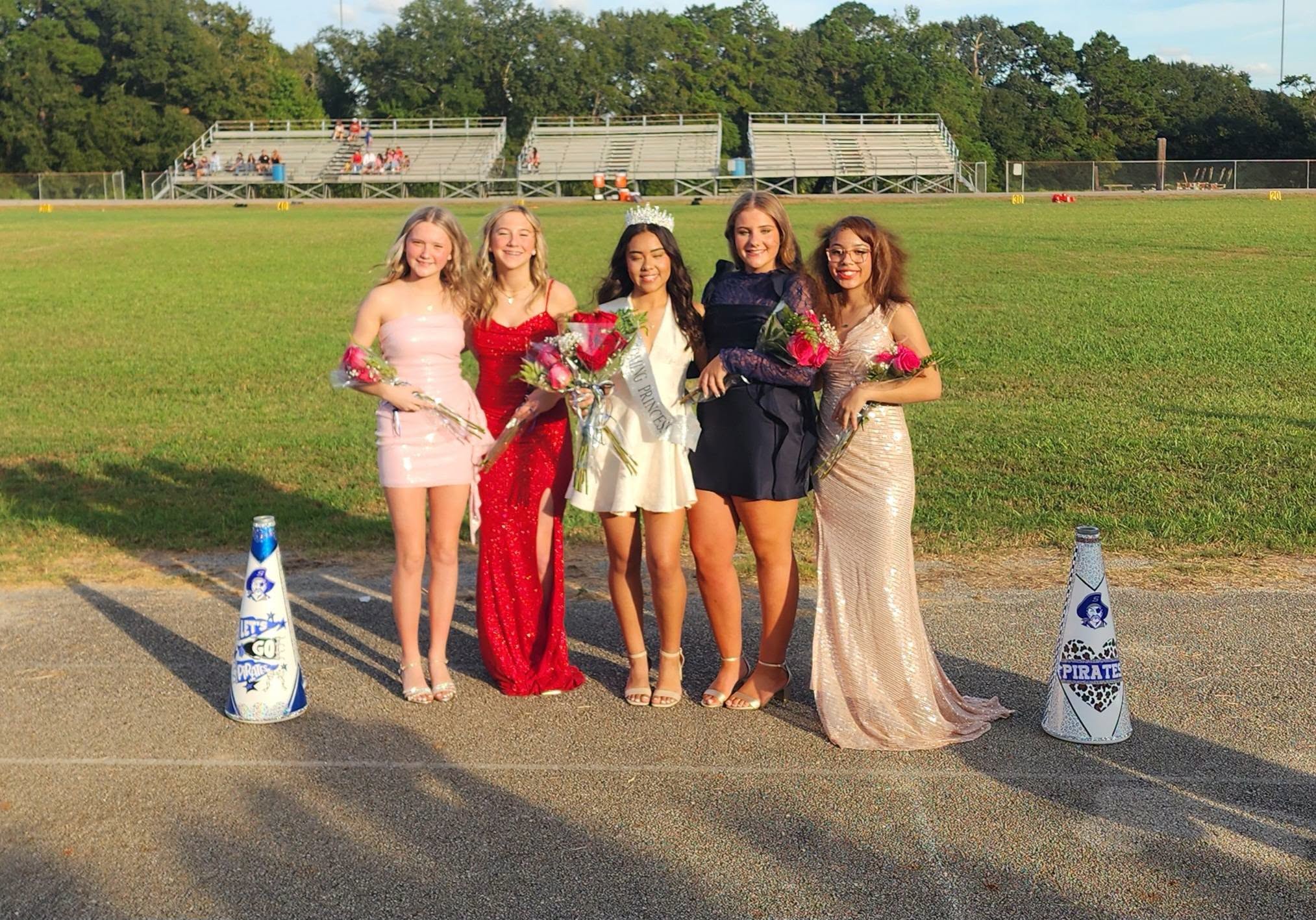 Five women stand on a track, each holding flowers, with a grassy field and bleachers in the background.
