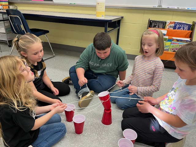 Students balancing cup onto of another cup while using teamwork and strings