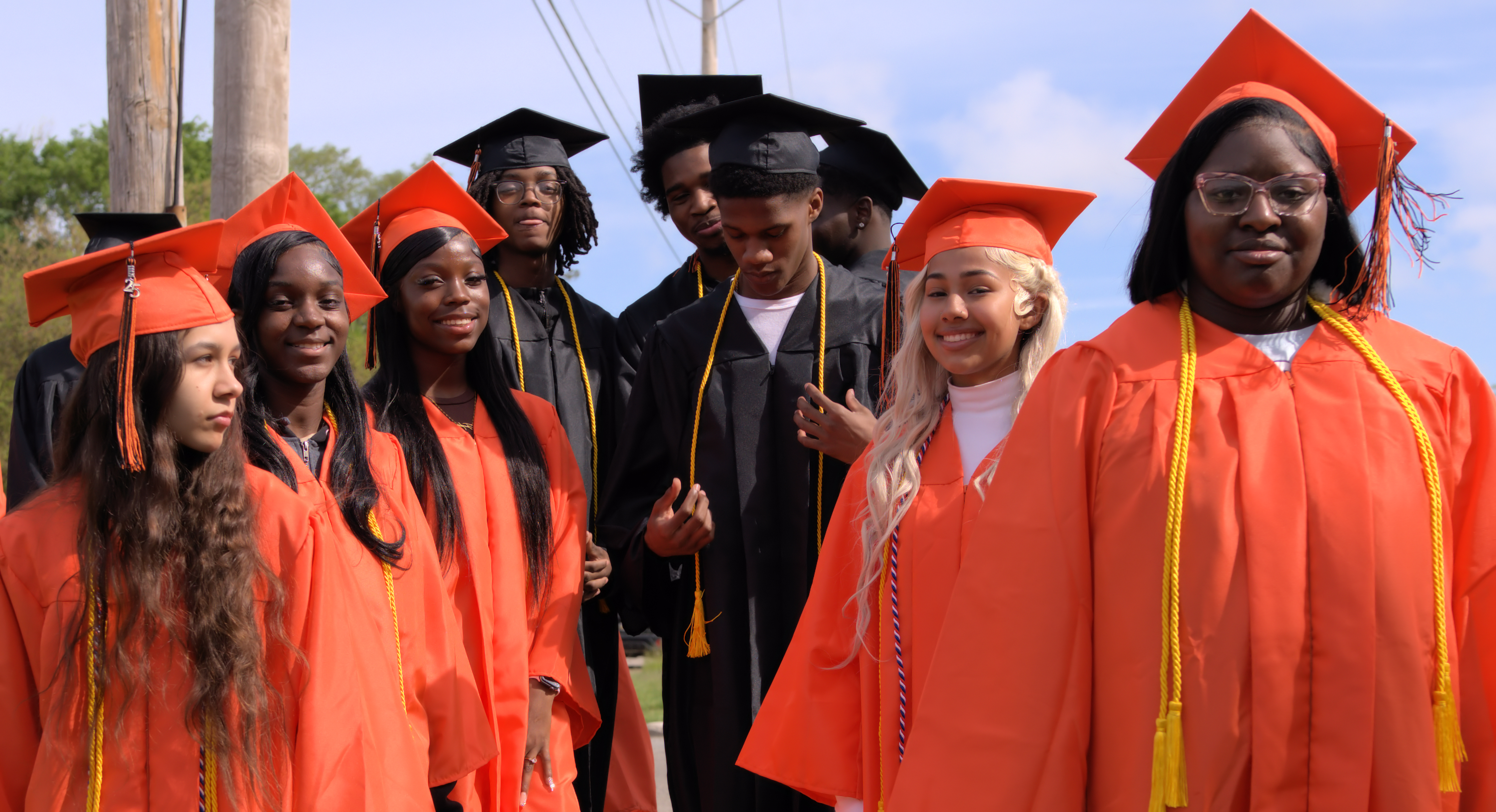 Group of high school graduates in orange and black caps and gowns standing together outdoors before the graduation ceremony.