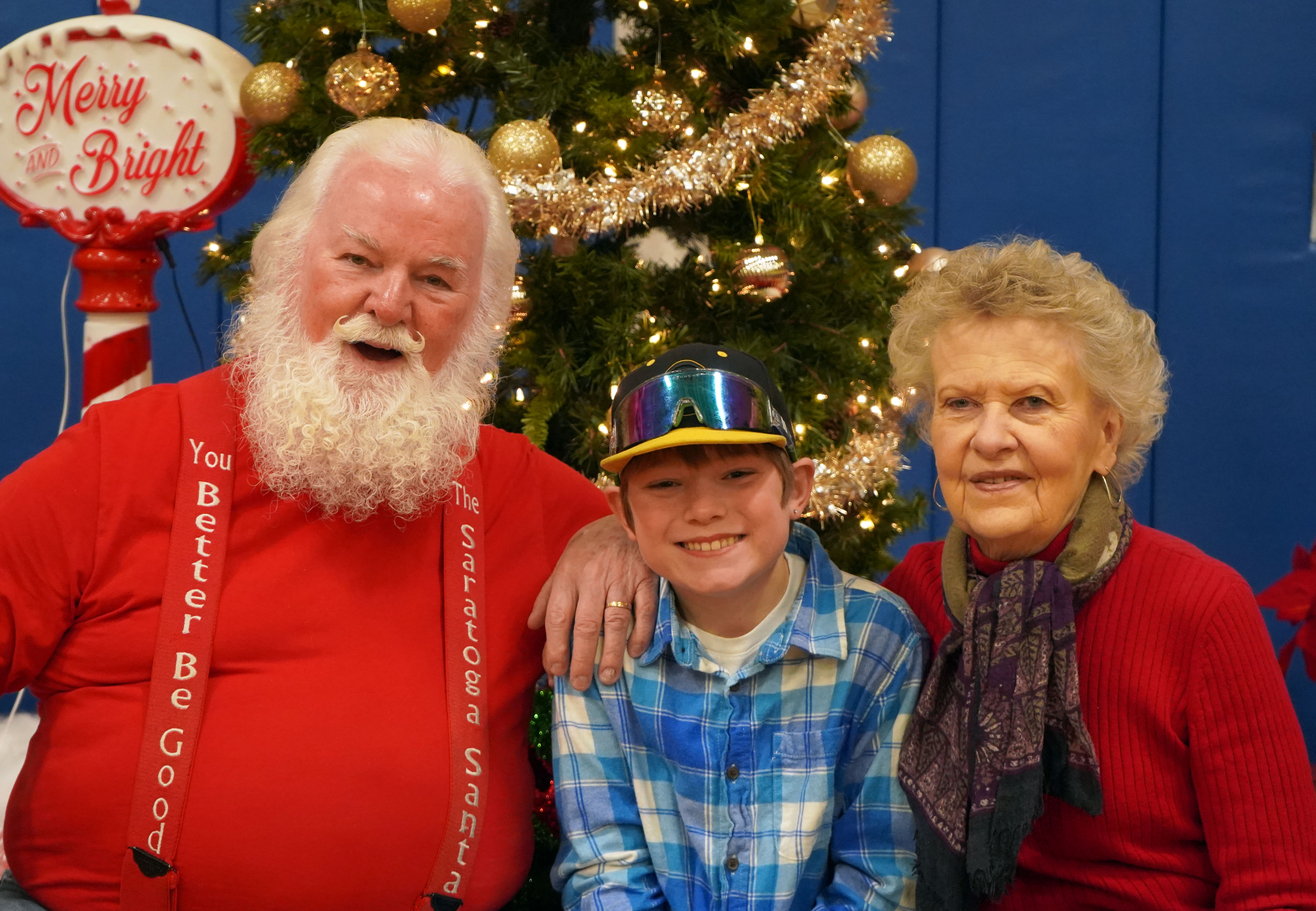 Child sits in front of Christmas tree with grandparents, one of whom looks like Santa.