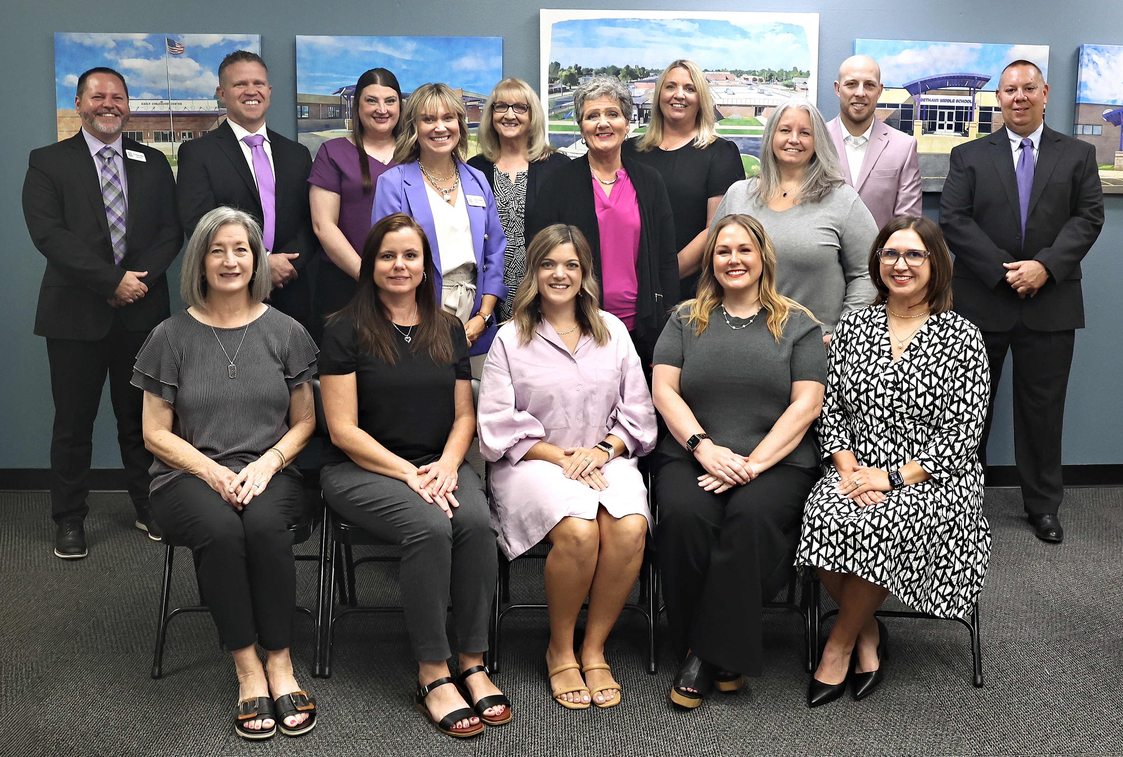 Group photo of Bethany Public Schools administrators and staff. Four women are seated in front and eleven staff members stand behind them. Several people are wearing shades of purple, gray, and black. The group is posed against a blue-gray wall with framed photos of school buildings in the background. Everyone is smiling toward the camera.