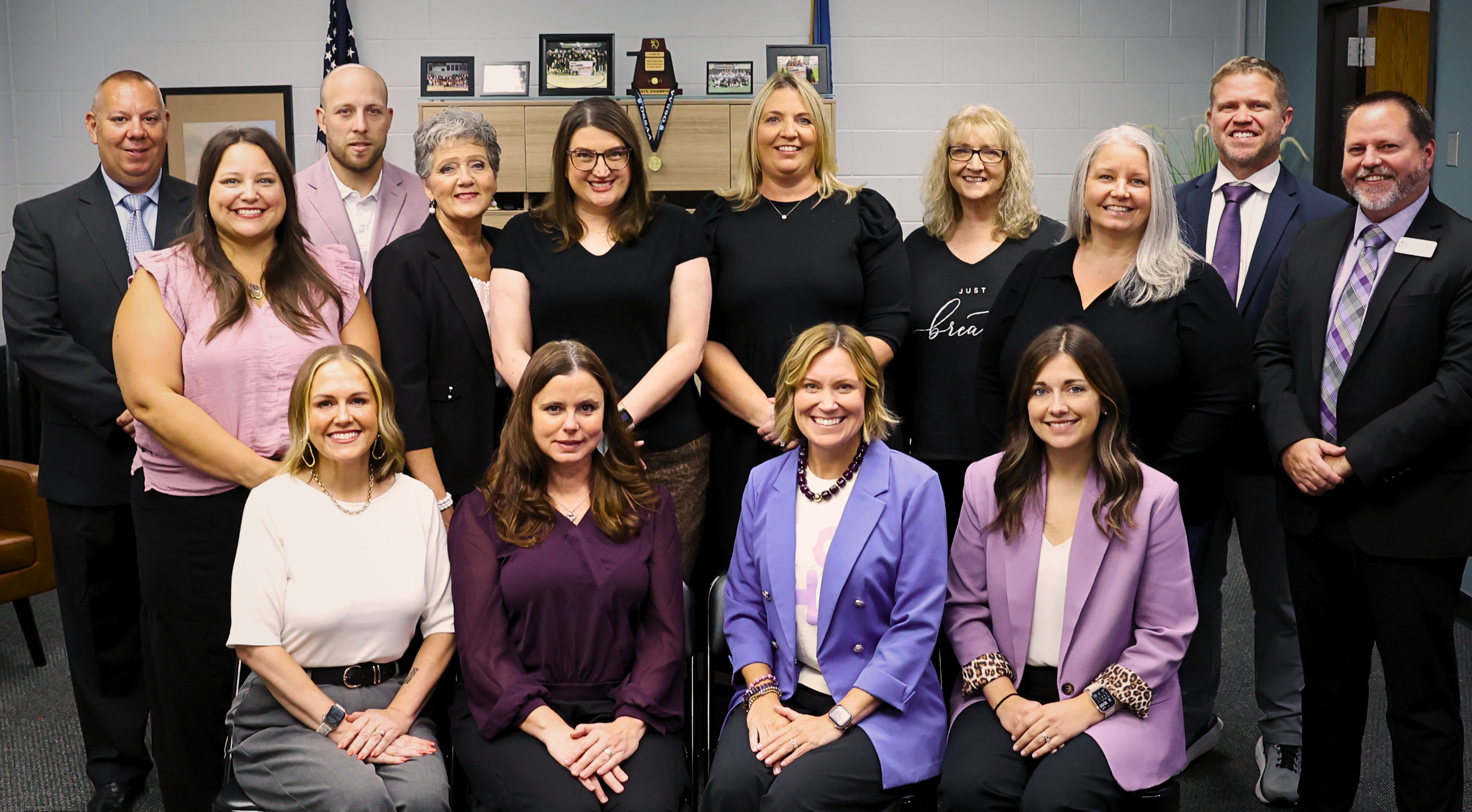 Fifteen Bethany Public Schools administrators smile for a group photo in the superintendent’s office, dressed in professional attire featuring shades of purple, black, pink and white. American and Oklahoma flags, framed photos and school awards are displayed in the background.