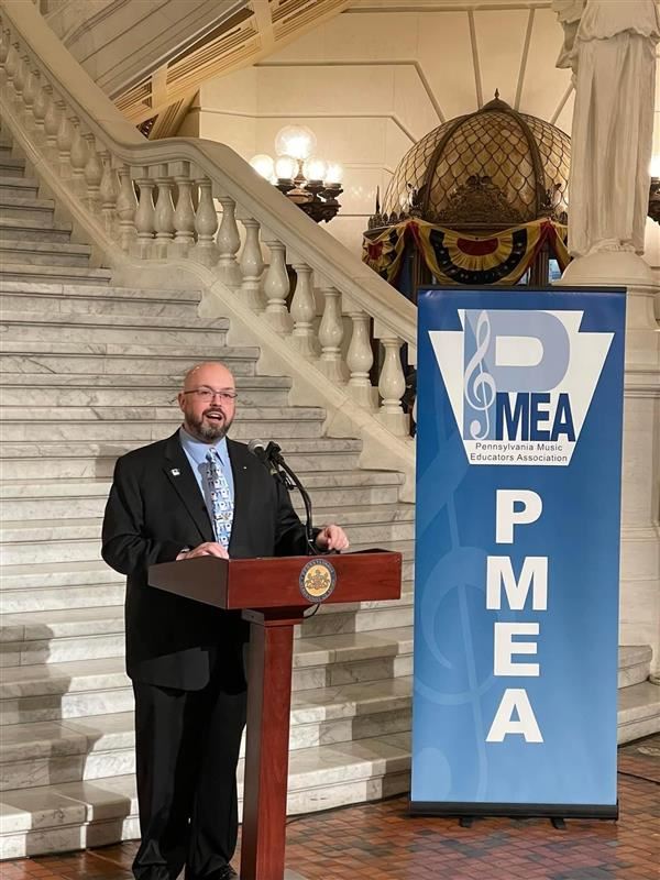A man in a suit standing at a podium, speaking at an event, with a sign displaying "PMEA" and "PMA".