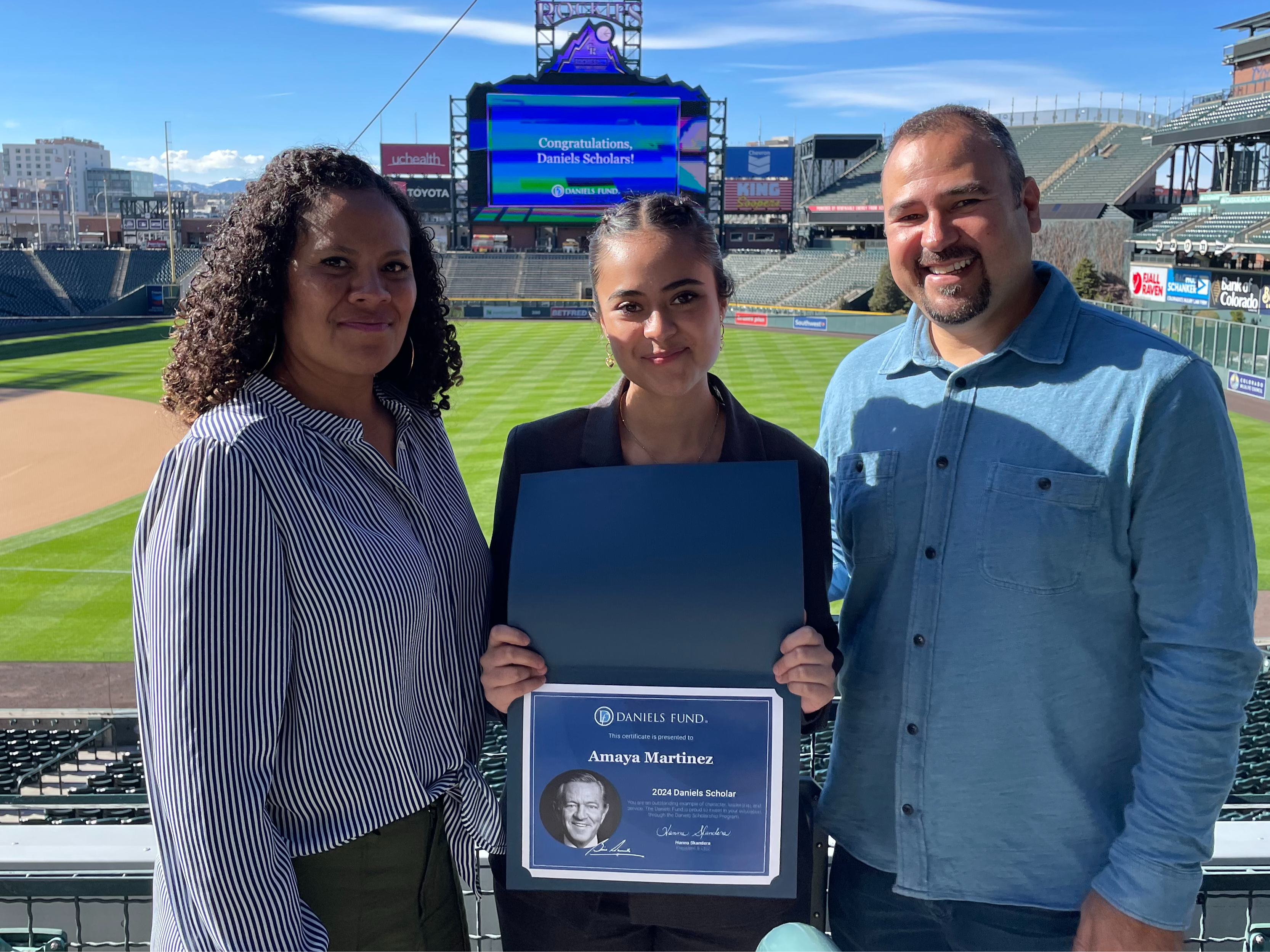 Amaya Martinez, a DPS student, receiving  an award for being a Daniels Fund Scholar. Amaya is posing with her parents from the stands behind the first base dugout at Coors Field. 