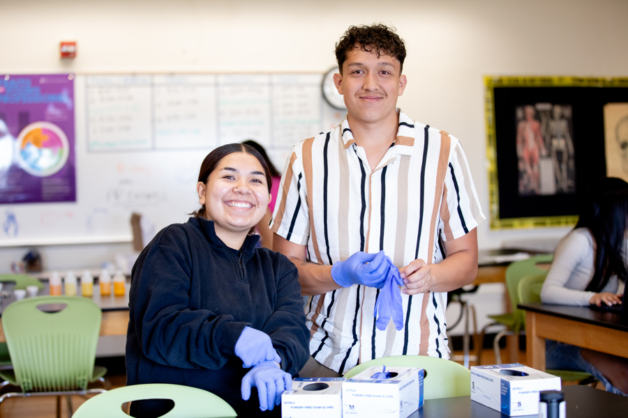two biomedical science students smiling and posing. The male student is wearing a striped shirt and the female student is wearing a black top. They both have gloves on. 
