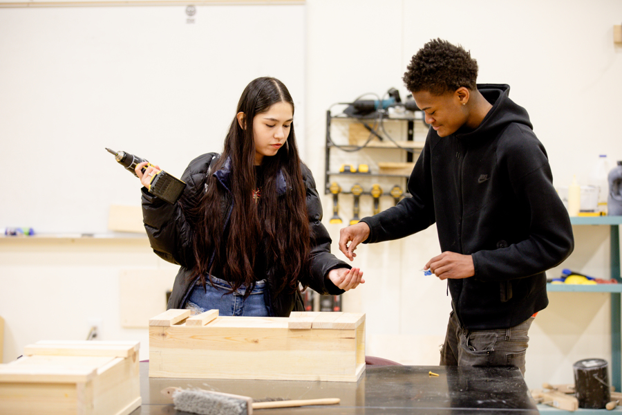 2 students work together in construction class. Male student is handing female student small screws while she is holding a power drill