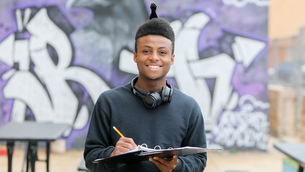 Young man holding a notebook in front of a wall with a graffiti